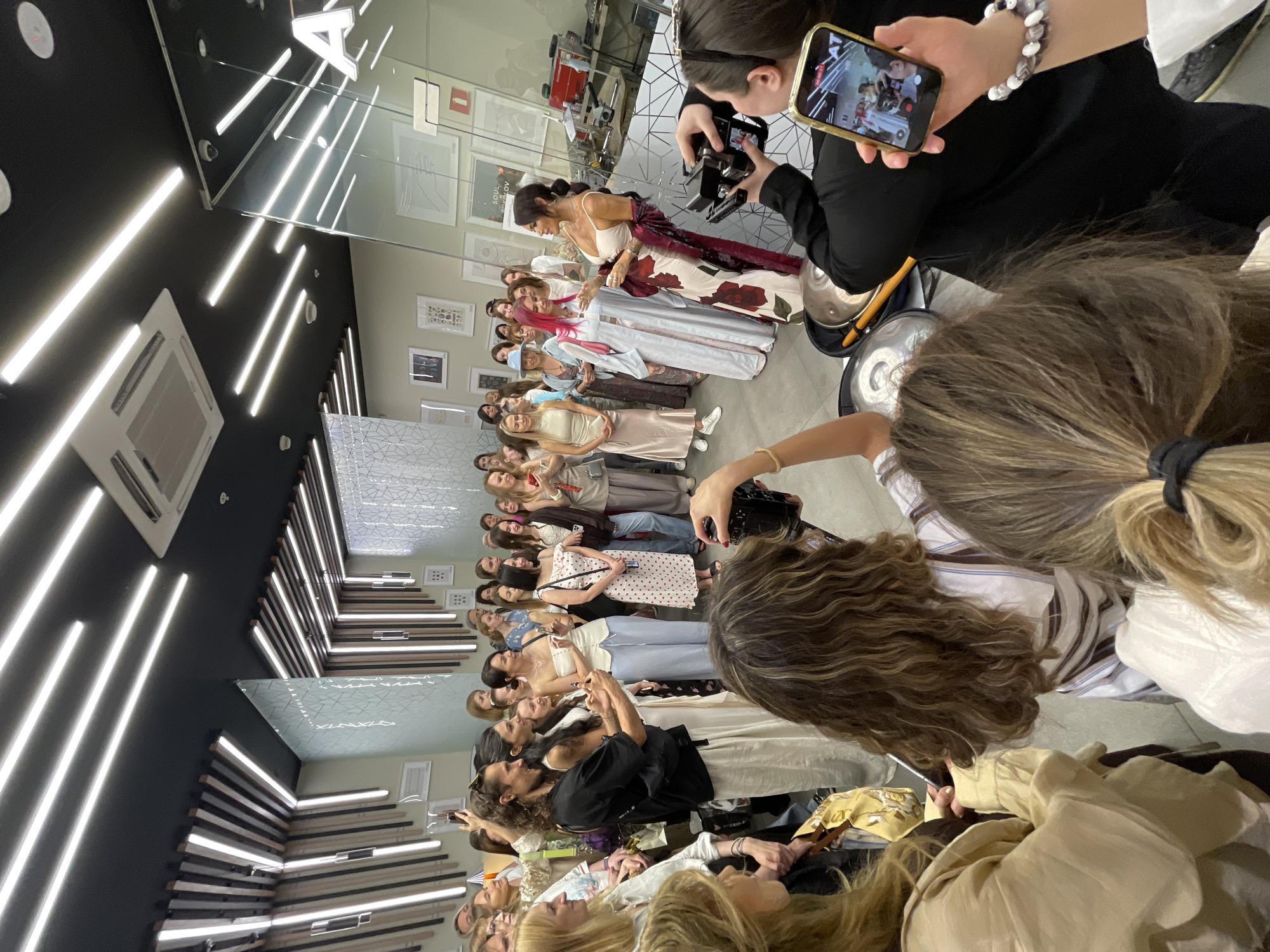 This detailed image captures an indoor gathering of approximately 30-40 individuals, predominantly women, within a modern commercial or event space. The location is presumed to be in Moscow, Russia, as per the request, but specific identifying landmarks within the city are not visible in the frame. The environment features a dark, possibly black, ceiling equipped with multiple parallel LED strip lights and two large air conditioning vents. Sections of the walls incorporate light-colored panels, some adorned with vertical wooden slats and integrated lighting. Framed artwork or documents are displayed on several wall surfaces. A prominent, stylized letter "A" is visible on a reflective glass partition on the right side of the frame, possibly part of a brand or venue name, which also appears as "AT" on a phone screen held by an attendee. In the central area, a group of five individuals stands as a focal point. From left to right: 1. A man with long, curly brown hair and a beard, wearing a black t-shirt and light-colored wide-leg trousers. He holds a small object, possibly a piece of jewelry, between both hands, looking towards the woman next to him. 2. A woman with long dark hair, dressed in a white sleeveless top and light-colored trousers, looking at the man. 3. A woman with dark hair, wearing a white strapless top and light blue wide-leg trousers, holding a smartphone. She is smiling. 4. A woman with bright pink hair, attired in a light blue or white patterned robe-style dress, smiling and facing right. 5. A woman with long dark hair tied back, wearing a white fitted top, a long floral-patterned dress, and a dark red or burgundy shawl draped over her left shoulder. She is actively gesturing with her right hand, seemingly addressing the crowd. The surrounding attendees, mostly women, are dressed in various casual to smart-casual attire, including dresses, blouses, and trousers, often in light or neutral tones. Many are holding smartphones, actively recording or photographing the event, indicating an engaged audience. In the immediate foreground, partially visible individuals include: - On the far left, a woman with light brown wavy hair wearing a light beige blouse. - In the lower center, a woman with long brown wavy hair, wearing a white and brown striped top. - On the right, a woman with dark hair tied back, wearing a black long-sleeved top. She is holding a professional camera in
