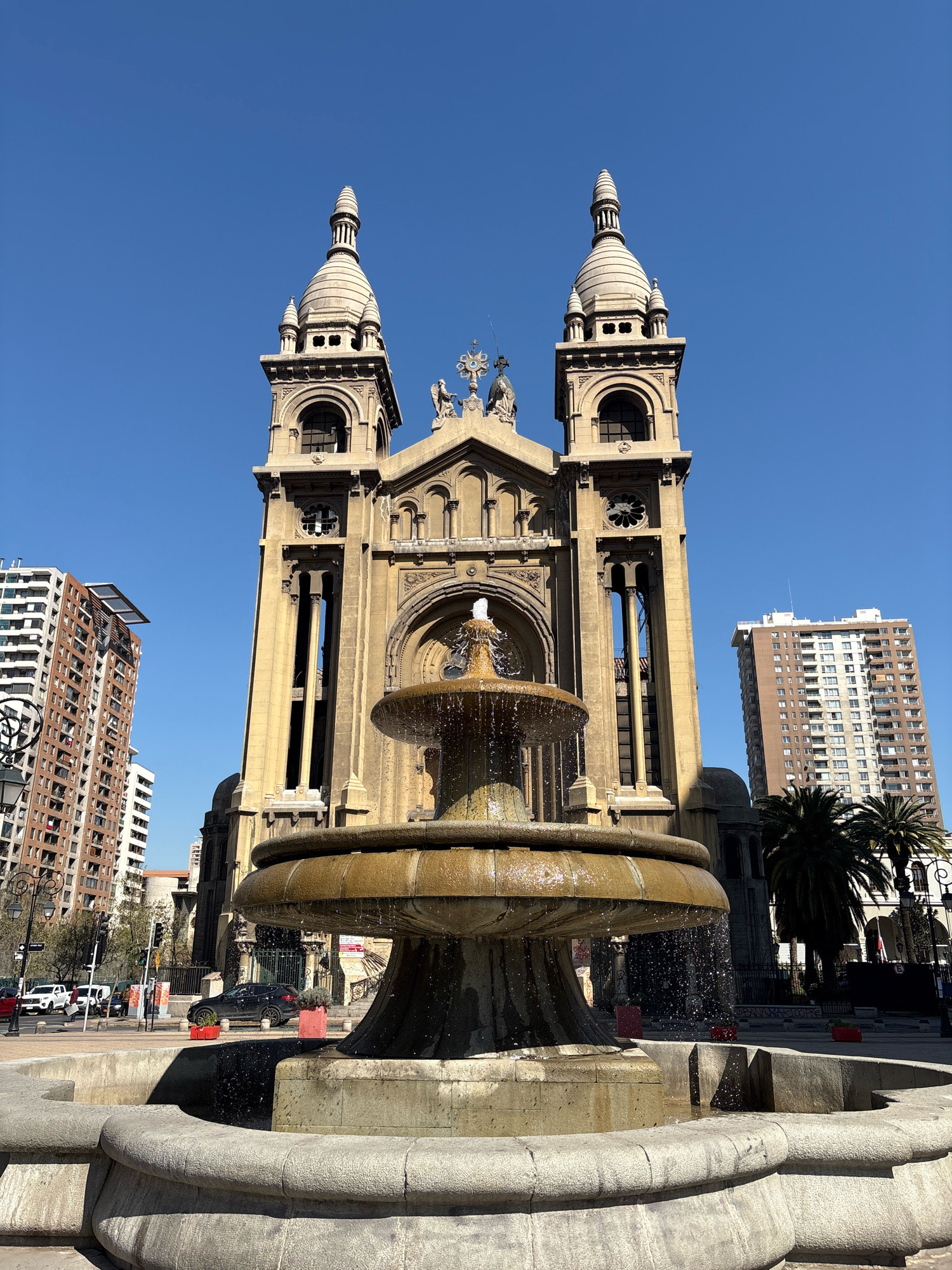 The image depicts the Metropolitan Cathedral of Santiago, Chile, with a large stone fountain in the foreground. The cathedral is a prominent, light-brown stone building with two bell towers, each topped with a spire. Sculptures of angels are visible on the roofline between the towers. A fountain with multiple tiers is actively spraying water, creating a misty effect. In the background, tall residential buildings with many windows are visible on either side of the cathedral, indicating an urban setting. The sky is clear and blue, and the sun is casting shadows. Cars and streetlights are present in the street area between the fountain and the buildings.