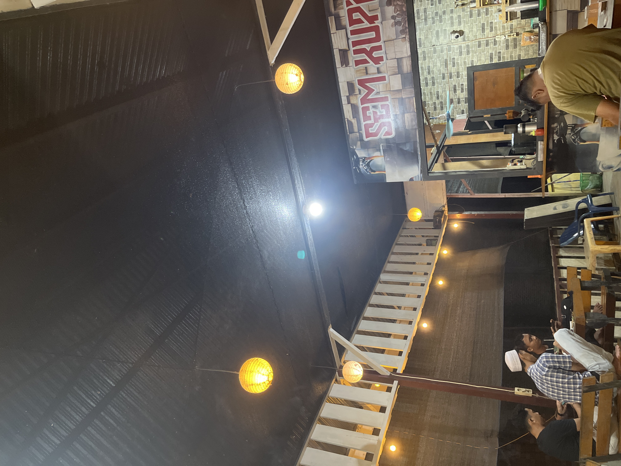 An overhead, slightly angled shot captures the interior of a dimly lit establishment, likely a cafe or restaurant, in Keudematangkuli, Indonesia, during the evening or night. The main light sources are several spherical, yellow-orange lanterns and some brighter, bare bulbs, casting a warm glow.
On the left, a dark, corrugated ceiling stretches across the upper half of the image. On the right, a prominent white structure resembling a staircase or tiered seating area descends into the lower part of the frame, also illuminated by hanging lights.
In the upper right quadrant, a person wearing a light brown/olive green shirt is leaning over a counter, seemingly engaged in preparing something, with various equipment visible on the surface. Behind this counter is a wall with a gray brick pattern. Above the counter, a large sign with red lettering on a wood-block-patterned background is displayed, showing text that appears to read "SEM-KOPZ" or "SEM-KOPI," suggesting a coffee-related business.
In the lower right, below the white tiered structure, several patrons are visible. One man in a blue plaid shirt and a white skullcap (kufi) looks upwards, while another man in a dark t-shirt is positioned below him. They appear to be relaxing or socializing in a seating area adjacent to the stairs. The overall atmosphere is intimate and relaxed, characterized by the soft lighting and the arrangement of space.