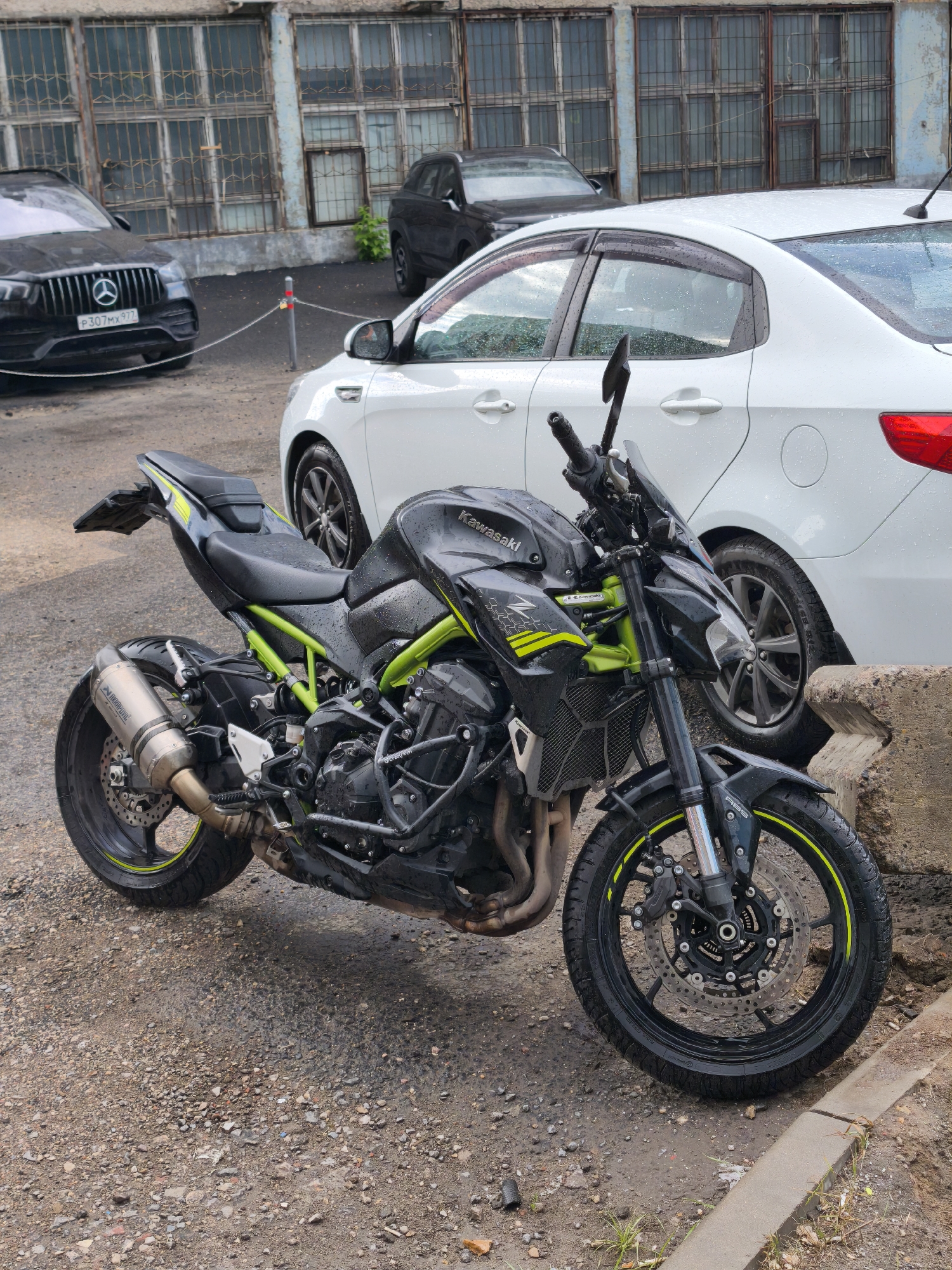 A black and green Kawasaki motorcycle is parked on a wet, pebbled surface, facing right. The motorcycle features an Akrapovic exhaust, neon green framework, and a wet seat. Behind the motorcycle, a white Kia sedan is parked, partially obscured by the motorcycle. A black Mercedes SUV is visible in the background to the left, with a license plate beginning with "P307". A dark-colored SUV is also visible further back, behind the white car. In the backdrop, a weathered building with multiple barred windows is present. This scene is most likely located in the city of Moscow, Russia, due to the license plate style.
