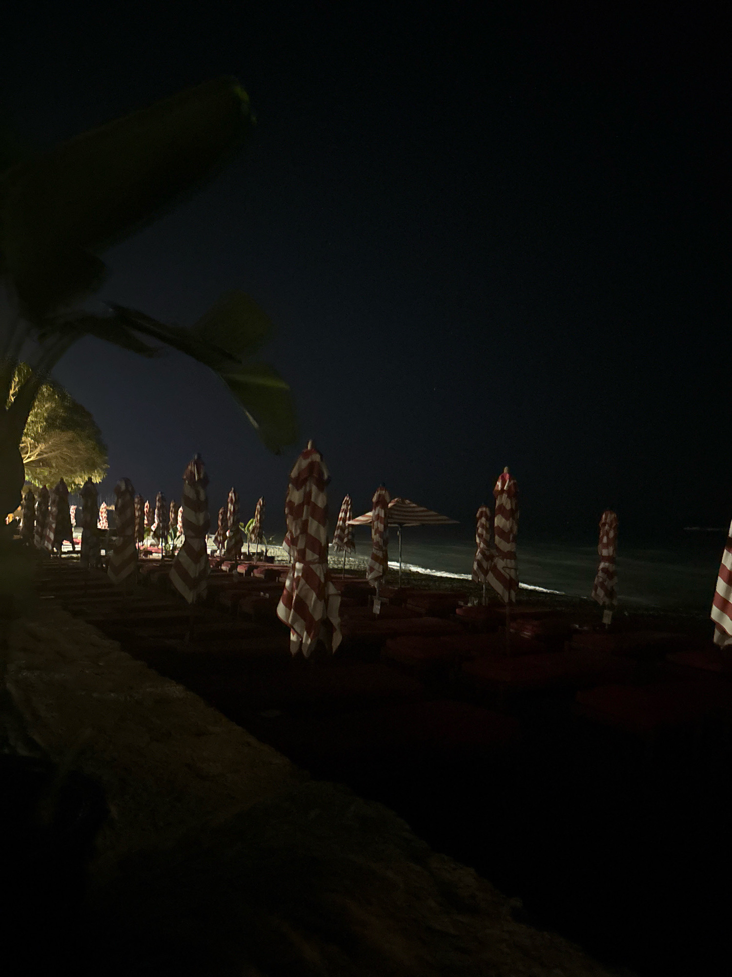 The image depicts a beach at night in the Agios Tychona Community, Cyprus. The scene is dimly lit, with a dark, star-filled sky above. Rows of closed beach umbrellas with red and white stripes are visible, set against the dark sand. Some umbrellas are folded, while others are partially open. In the foreground, the sand is textured and appears damp in places. To the left, the silhouette of a large tree with sparse foliage is prominent, casting shadows. The sea is visible in the background as a dark, indistinct line. The overall atmosphere is one of quietude and stillness, typical of a beach after closing hours.
