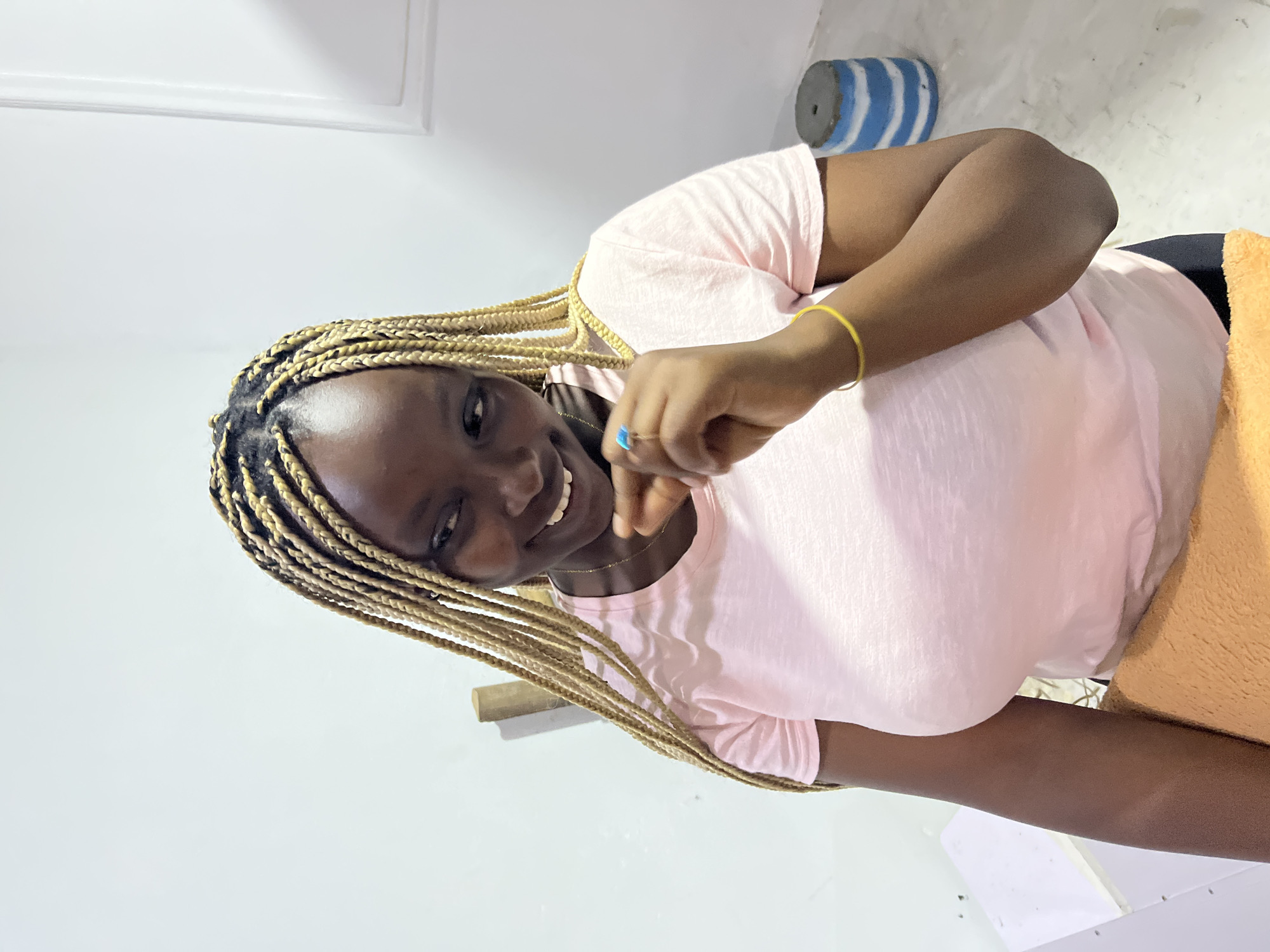 A dark-skinned woman with blonde braids poses indoors. She is wearing a pink t-shirt, a yellow bracelet, and a ring with a blue stone. Part of an orange blanket is visible on her right side. In the background are white walls and a cylindrical object with blue and white stripes. The location is an indoor space in the city of Unknown, Unknown.