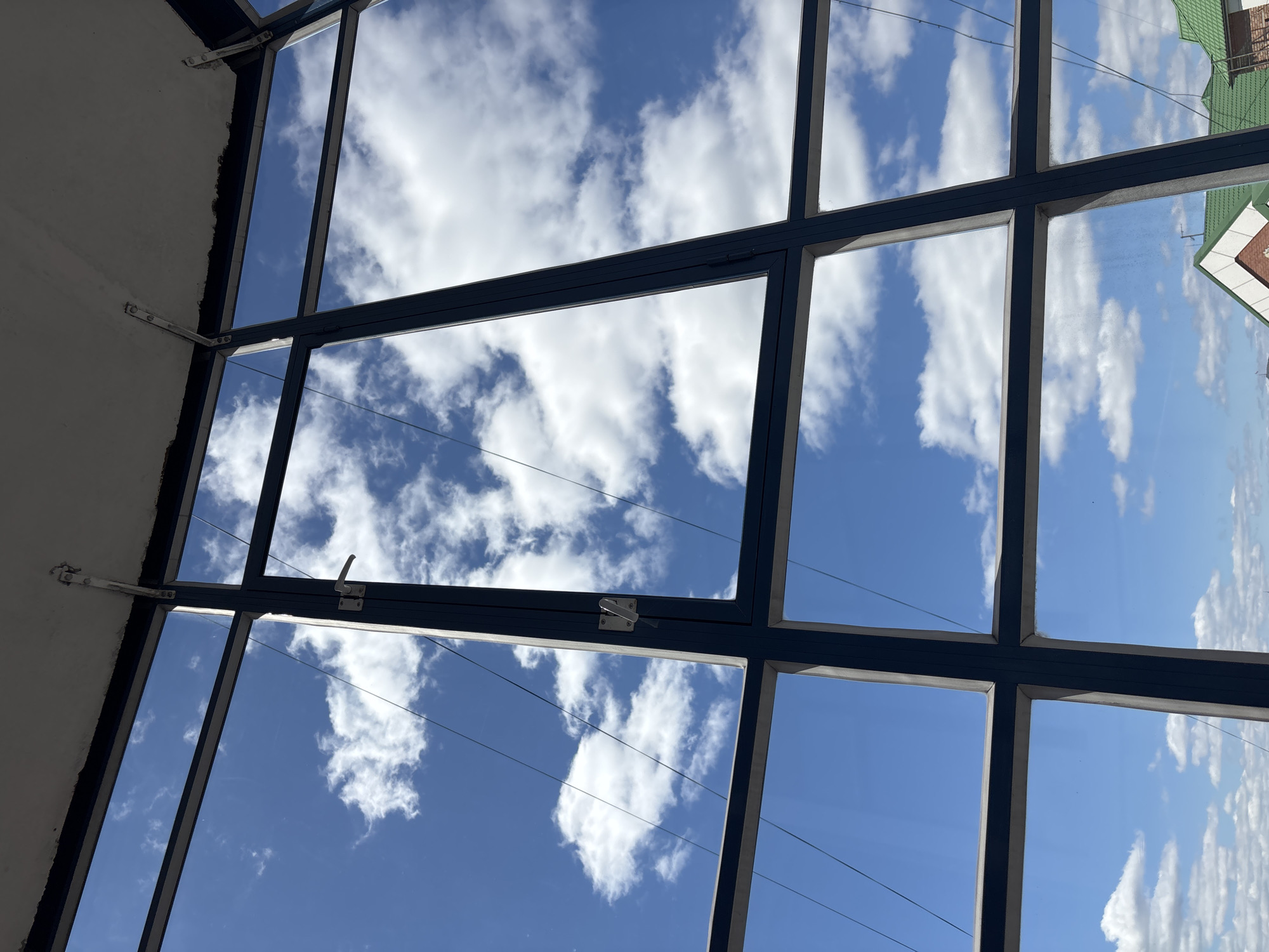 The image shows a window with a view of the sky with clouds. The window is made of dark metal frames that form a grid pattern. The view through the window is of a bright blue sky dotted with white, puffy clouds. The image is taken from inside, looking up and out through the window. Outside the window reflection a building is visible. The location is Krasnoyarsk, Russia.