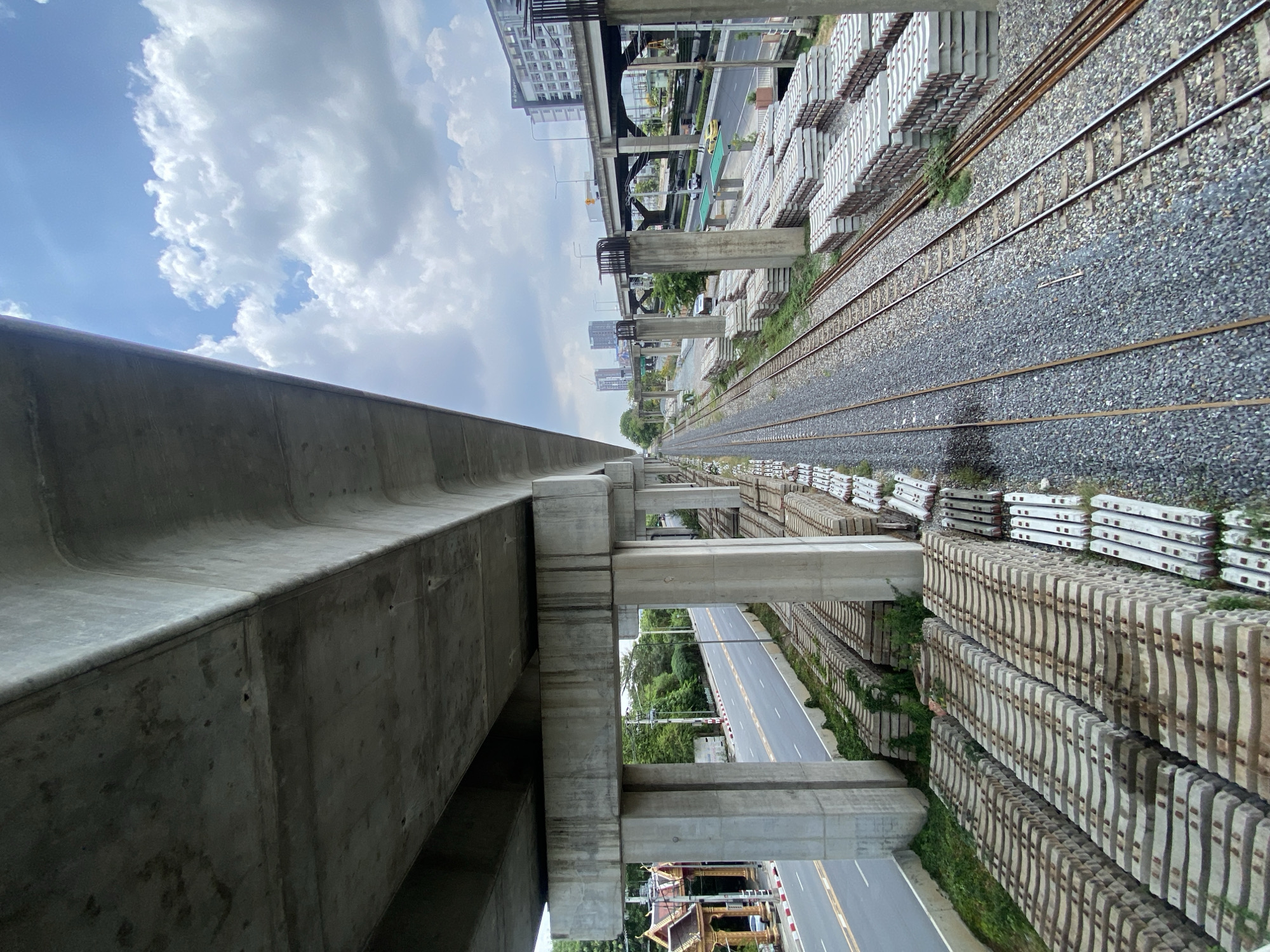 The image depicts a scene in Bangkok, Thailand, showing infrastructure and urban elements. A large concrete elevated structure dominates the left side of the frame, with support columns extending downward. Adjacent to this structure are elevated railway tracks, with gravel ballast and metal rails. Stacks of precast concrete elements, possibly railway sleepers, line both sides of the tracks. Below the elevated structures is a road with visible lane markings. Patches of green vegetation are interspersed among the concrete structures and along the roadside. In the distance, buildings and other infrastructure elements are visible, creating an urban backdrop under a cloudy sky.