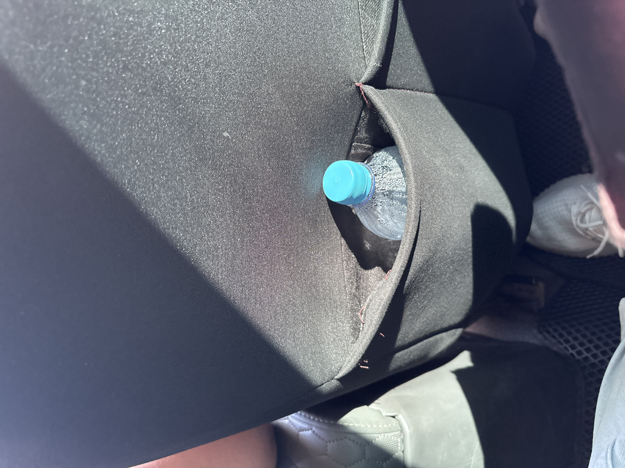 The image shows a close-up view of a black car seat organizer with a water bottle inside. The organizer is attached to the back of a car seat and has a pocket where the bottle is placed. The bottle has a blue cap and is partially filled with water. The car seat is upholstered in a dark fabric, and there is a shadow cast across it from a window. The background is out of focus, but it appears to be the interior of a car. The image does not contain any information that allows for the determination of its location in the city of Pavlodar, Kazakhstan.