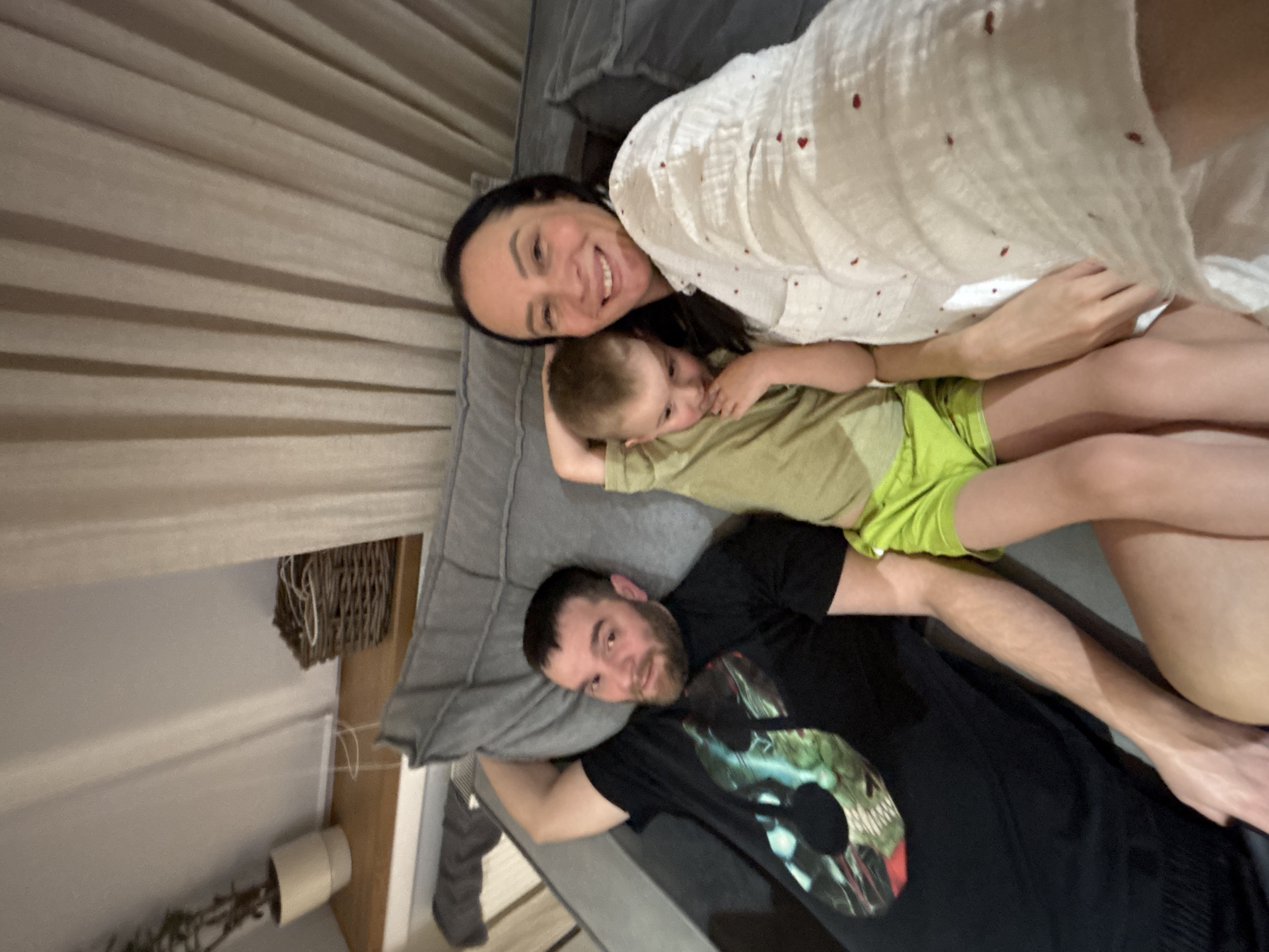 The image displays three individuals—a man, a young boy, and a woman—seated on a grey sofa in an indoor residential setting within the city of Chaikovsky, Russia. The man, positioned on the left, has short dark hair and a beard, wearing a black t-shirt with a complex graphic design featuring green and blue elements. He reclines with his right arm bent behind his head, looking towards the camera with a subtle smile. A young boy, with light brown hair, is seated between the man and the woman, wearing a light olive green short-sleeved top and bright lime green shorts. His right hand is near his mouth or chin, and he is looking slightly downwards. The woman, on the right, has dark hair and is wearing a white, loose-fitting top adorned with small red heart patterns. She is smiling and looking directly at the camera, suggesting she is holding the device for a selfie. In the background, light-colored curtains are visible, and to the left, a wooden shelf holds a cream-colored potted plant and a brown wicker basket.
