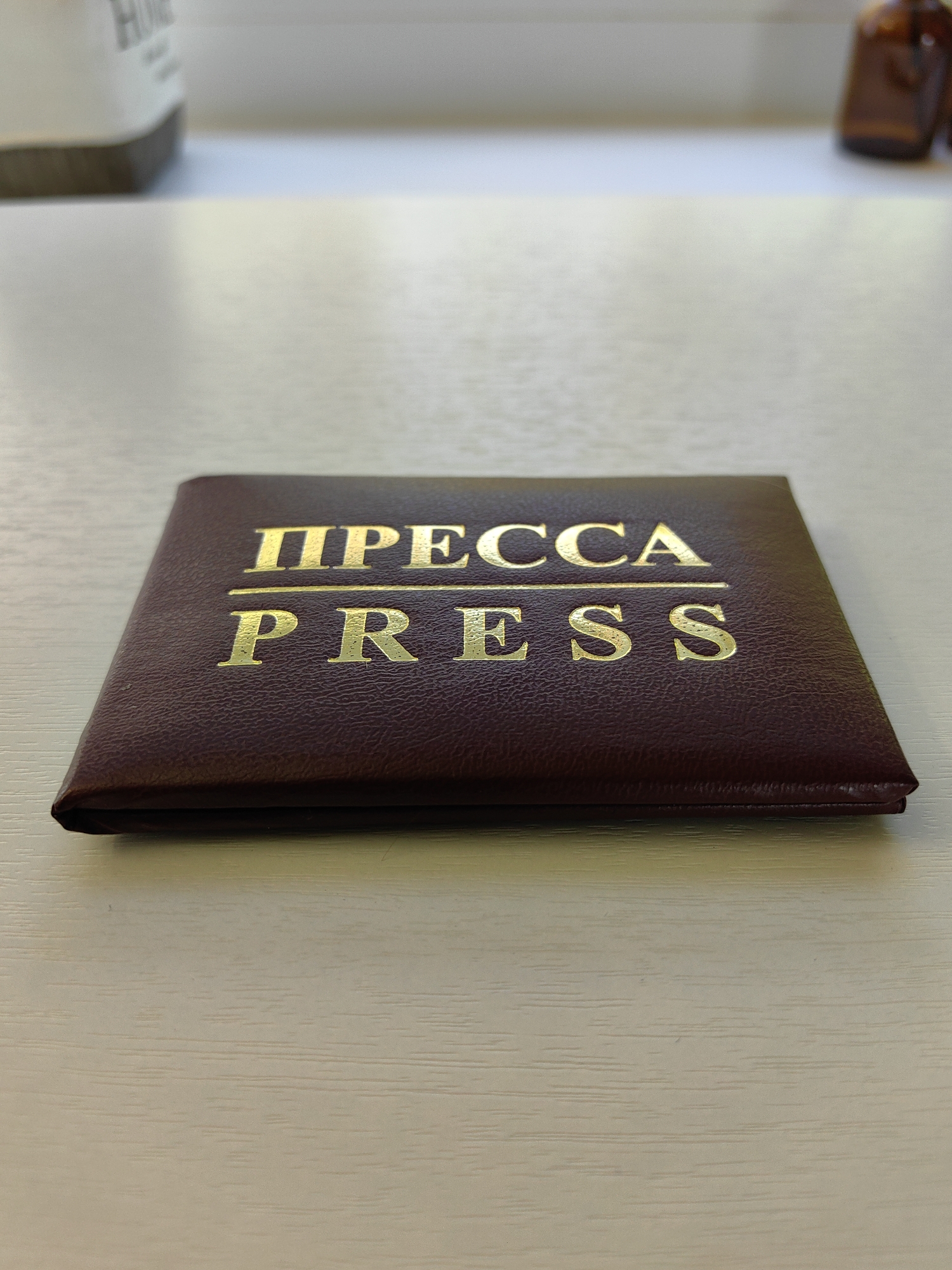 A close-up, eye-level shot captures a dark red, leather-bound pass with gold lettering on a light-colored, textured surface. The pass is positioned horizontally and slightly angled towards the viewer's right, with its spine facing the bottom edge of the frame. The gold lettering on the pass reads "ПРЕССА" on the top line and "PRESS" on the bottom line, separated by a thin gold line. The surface appears to be a wooden table or desk, with visible wood grain running horizontally across the frame. In the background, slightly out of focus, a white object with dark lettering is visible on the left, and two small brown glass bottles are visible on the right, suggesting an indoor setting. The lighting is soft and diffused, creating subtle shadows and highlighting the texture of the leather and the shine of the gold lettering.