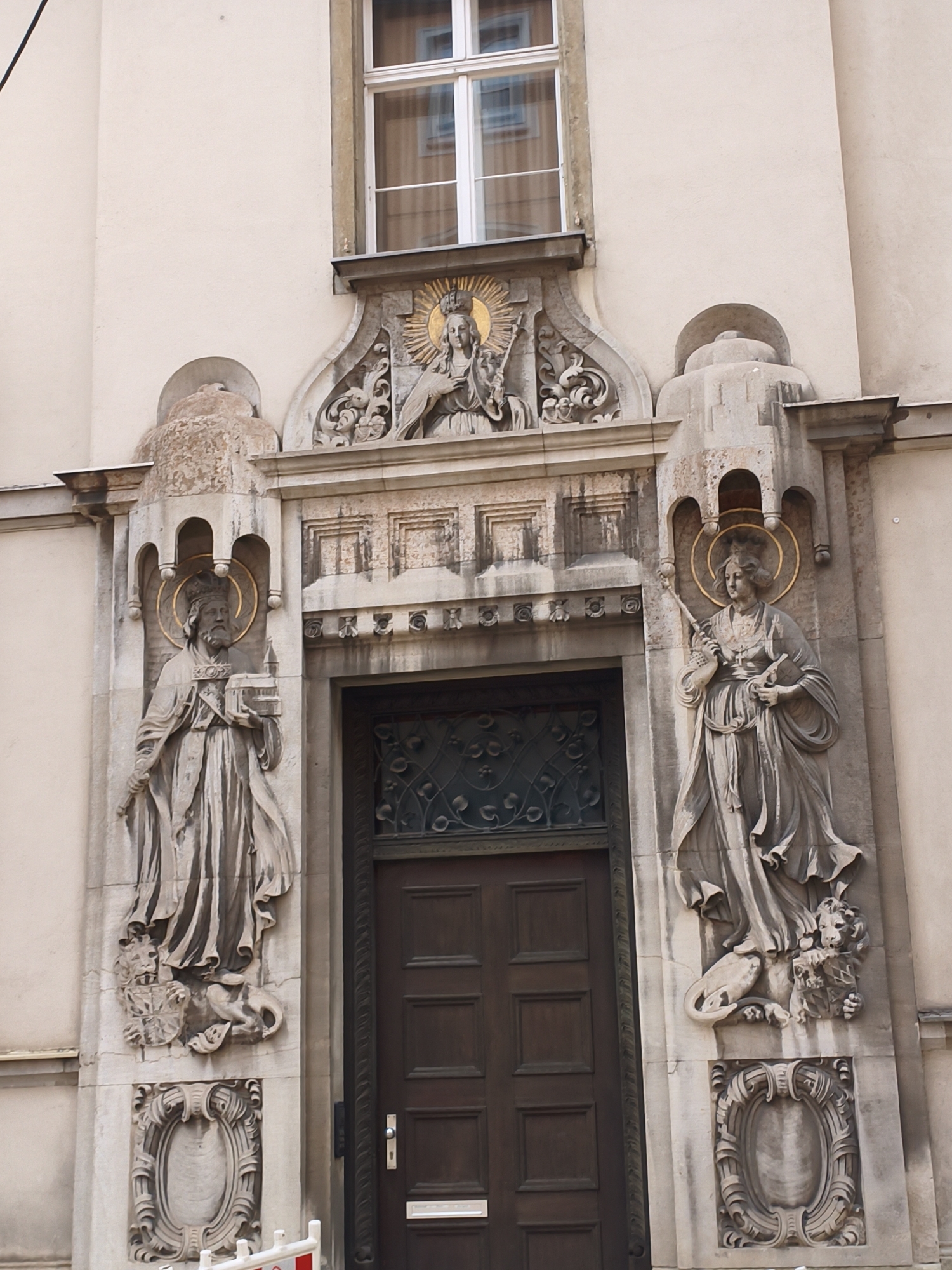 The image shows the facade of a building with a prominent entrance. Above the dark wooden door, which features decorative metalwork on the upper part, is a stone entablature with carved embellishments. Flanking the entrance are two stone sculptures of figures in robes, each with a halo. The sculpture on the left depicts a bearded male figure holding a model of a building and a staff, with a lion at his feet. The sculpture on the right shows a female figure holding a staff and a book. Above these sculptures, an ornate stone frame surrounds a relief carving of a crowned figure holding a child. A window with white framing is visible above this relief, set within a larger architectural element. The building's exterior is light-colored and textured. Decorative stone carvings, including cartouches, are present on either side of the entrance, below the full-length sculptures. The image appears to be taken from a street-level perspective, with part of a railing or barrier visible in the lower foreground. This is the facade of the Neupfarrkirche in Regensburg, Germany.