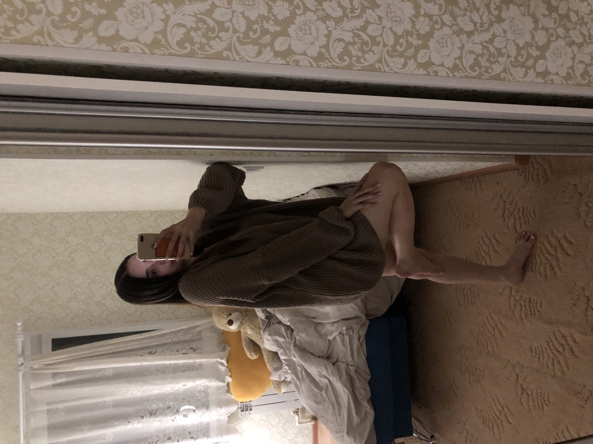 A woman sits on a bed in a room with floral wallpaper, visible in a mirror. The mirror shows a partial reflection of a window with white curtains, a radiator, a brown sweater on the woman, and a teddy bear. The woman holds a smartphone, presumably taking a selfie. A carpet covers the floor. It is uncertain if the location is Noviy, Russia.