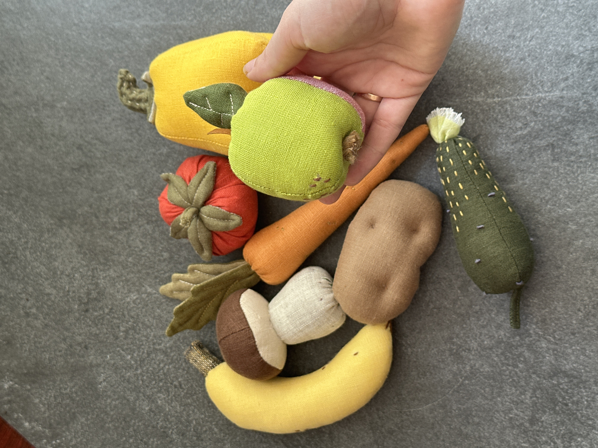 A flat lay of handmade fabric toy vegetables and fruit is displayed on a dark gray textured surface, likely a countertop or table. The collection includes a yellow banana, a brown and white mushroom, a brown potato, an orange carrot, a red tomato, a yellow pumpkin, a green pickle, and a green apple. A person's hand, wearing a gold ring on the right ring finger, is visible on the right side of the image, holding the green apple. The background is a plain, dark gray surface with a subtle texture. The scene is lit from above, casting soft shadows. The image is an artistic arrangement of fabric toys, with no indication of location in Chornomorske, Ukraine.