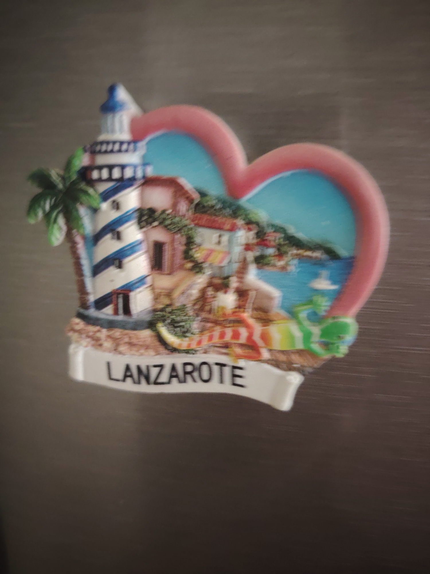 A heart-shaped magnet is attached to a metallic surface, likely a refrigerator. The magnet depicts a scene with a lighthouse, buildings, a palm tree, and a view of the sea with rolling hills in the background. A small green figure, possibly a lizard or frog, is positioned near the water. The word "LANZAROTE" is written in black capital letters on a white banner at the bottom of the magnet. The background is a blurred, neutral gray surface. The image quality is poor due to blurriness and low resolution. There is no information in the image to suggest a location in Huntingdonshire, United Kingdom.