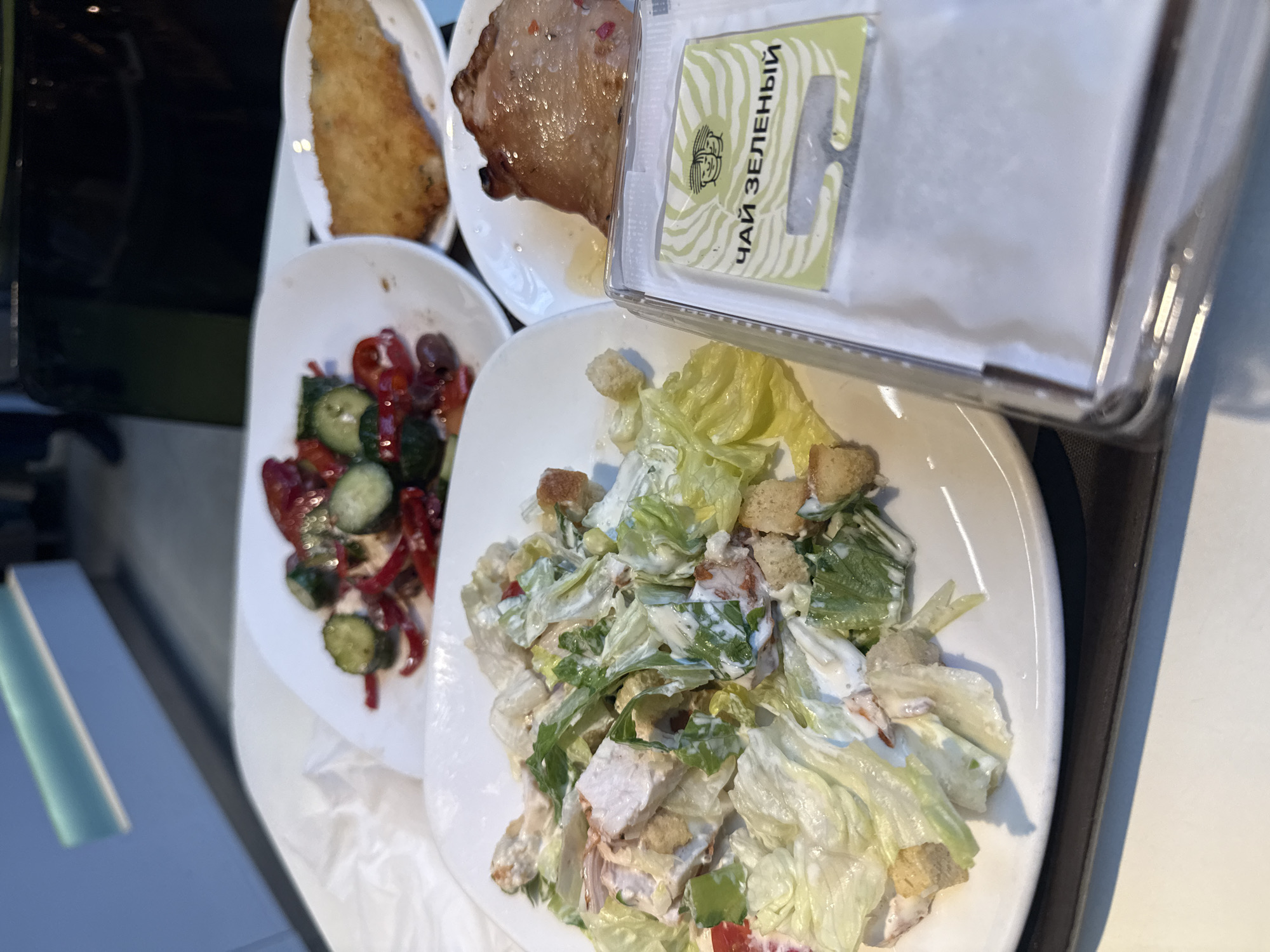 The image shows a tray of food, likely on an airplane, featuring a breaded fish filet, a cut of meat, a salad of tomatoes and cucumbers, and a Caesar salad with croutons. A small packet labeled "Чай Зеленый" (Green Tea) in Russian is also visible. The food presentation, tea branding, and overall setup suggest an airline meal service, but the specific location in Moscow, Russia, cannot be determined from the image alone.