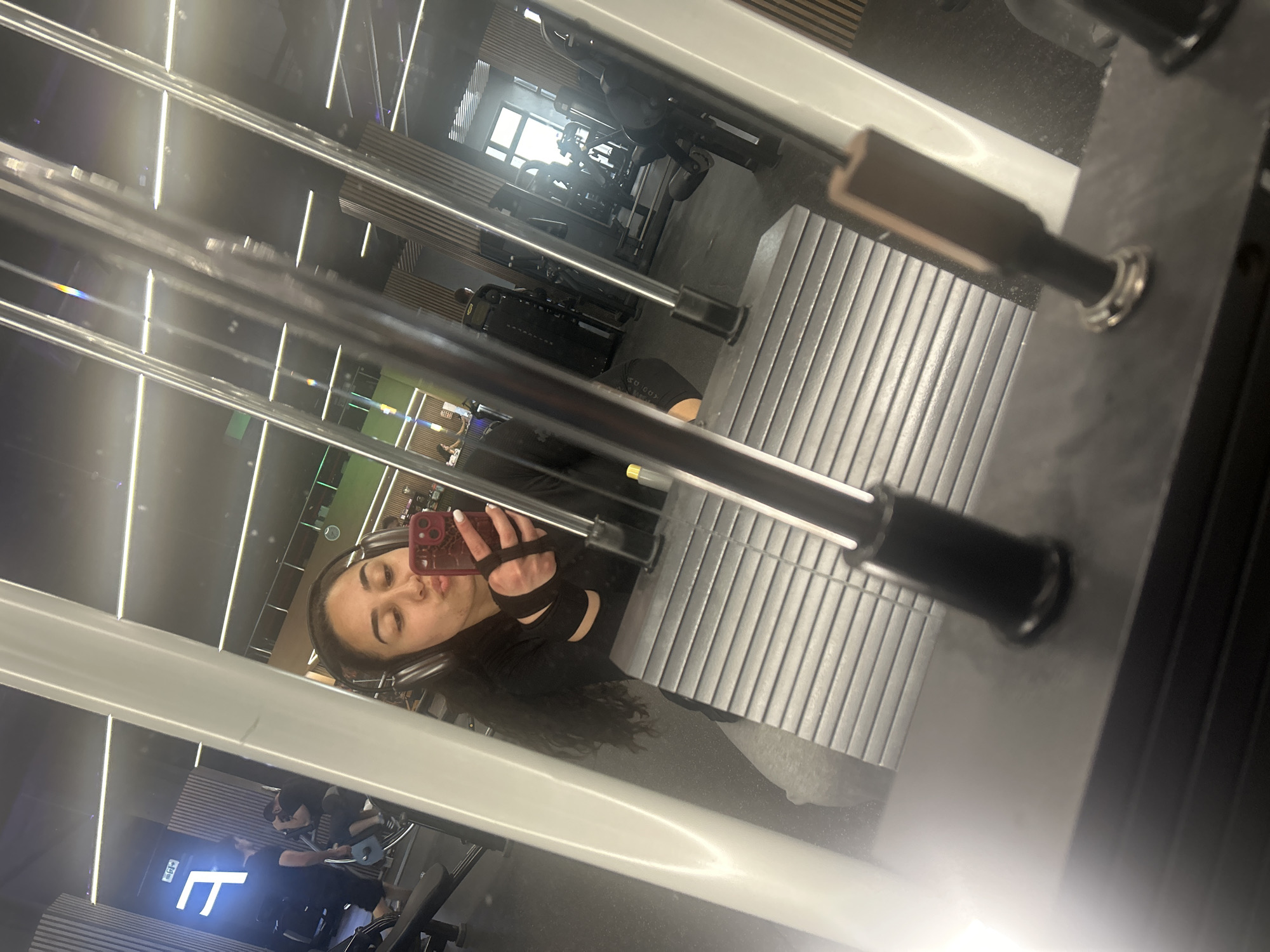 A reflection of a woman holding a phone in a gym's mirror. She has on headphones and is wearing black clothing. Gym equipment is visible in the reflection, including a weight machine. The gym appears modern with linear lighting fixtures. The location is likely Burgas, Bulgaria.