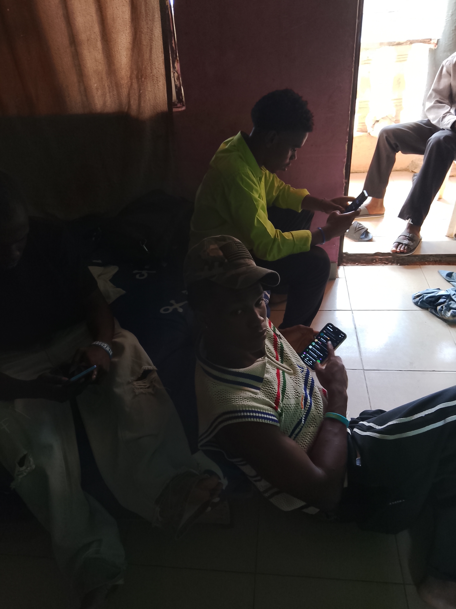 Three young men are indoors, likely in Nigeria, as suggested by the location context. The scene is dimly lit, with natural light entering from an open doorway at the far right, illuminating the floor and the legs of a man sitting on a stool. The men are casually dressed, with one wearing a bright yellow shirt, another in a white, blue, and red striped shirt and a cap, and the third in ripped light-colored jeans and a dark t-shirt.
All three men are engrossed in their mobile phones, holding them and looking at the screens. The man in the yellow shirt is seated on the floor and appears to be interacting with his phone. The man in the striped shirt is also sitting on the floor, holding his phone with his left hand and his right index finger touching the screen. The third man, partially visible on the left, is also looking at his phone, with his arm resting on his lap.
The setting appears to be a simple room with tiled flooring. A dark curtain hangs on the left side of the image, creating a strong contrast with the lighter area near the doorway. There are some dark items on the floor, possibly bags or clothing. The overall mood is relaxed, with the individuals absorbed in their personal digital activities. There is no indication of the time of day or weather outside. Visible text includes "NEW GROUP" on a bracelet worn by the man in the light-colored jeans.