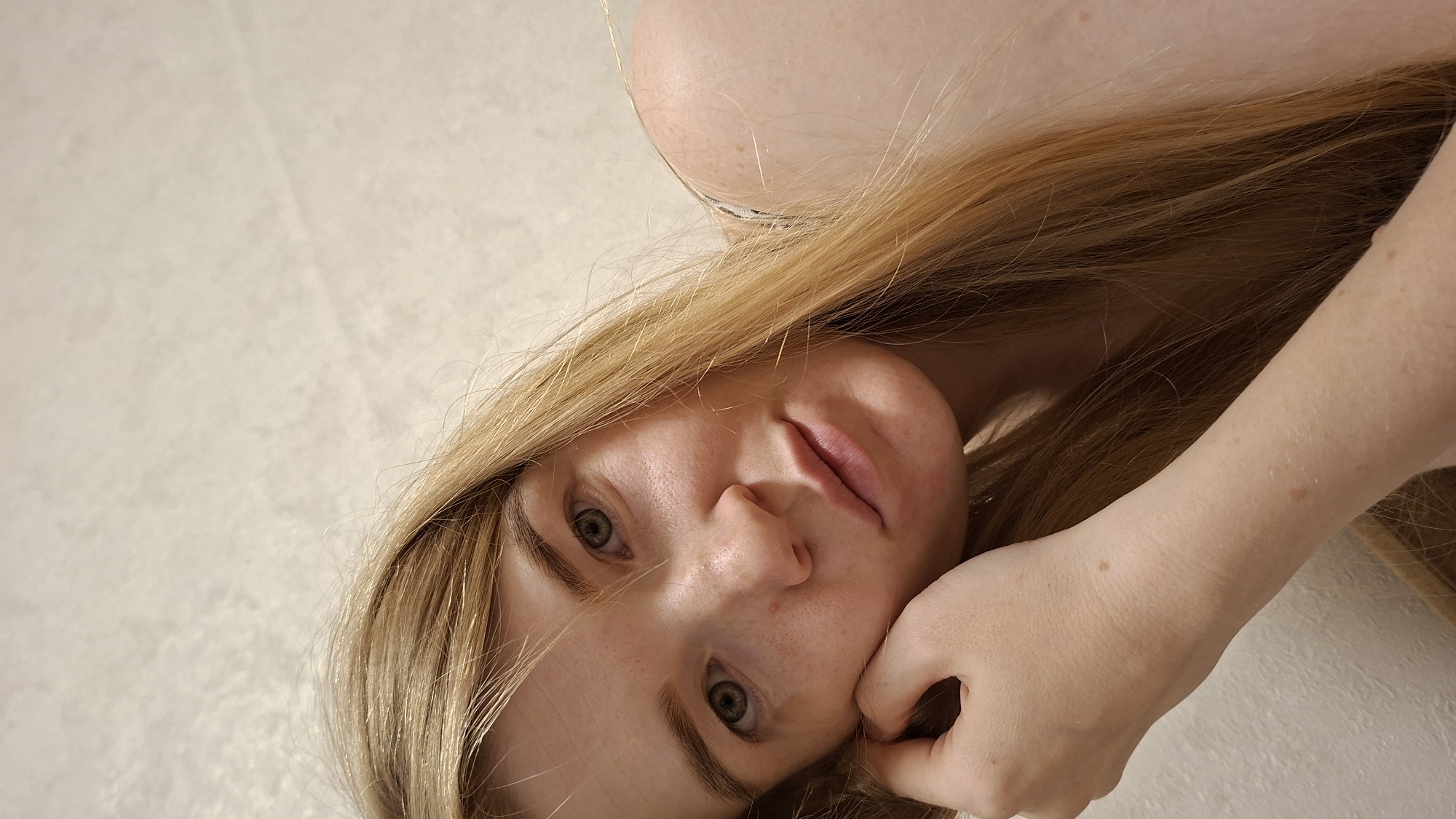 This is a close-up, sideways photograph of a person's face and arm. The subject has fair skin with freckles, long blonde hair, and green eyes. The person is looking towards the camera with a neutral expression, resting their head on their hand. The background appears to be a light-colored, textured surface, possibly a wall or floor. The lighting suggests it is daytime, with soft, diffused light illuminating the scene. There are no visible objects or text in the image. The context provided suggests the location is Bolshiye Bukory, Russia.