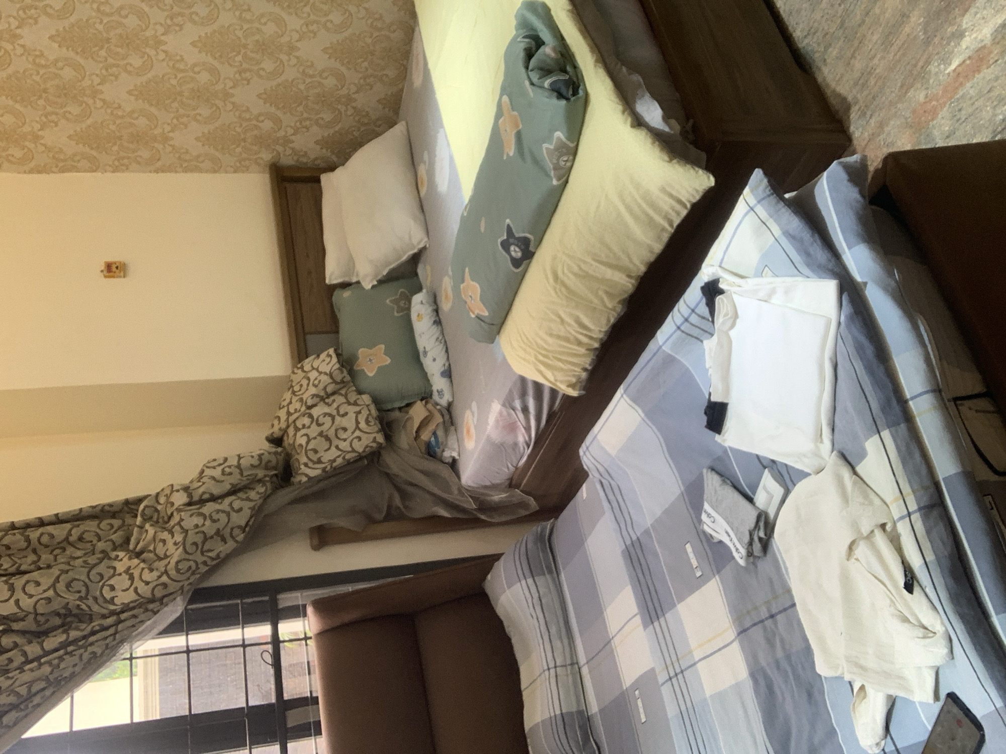 The image depicts a bedroom in Abuja, Nigeria. Two beds are visible, one with a brown upholstered headboard and the other with a wooden headboard. Both beds are made with patterned sheets and duvets. On the bed with the brown headboard, there are several folded clothes, including a white t-shirt and grey undergarments. On the other bed, there are pillows and a rolled-up duvet with a star pattern. A window with patterned curtains is to the left, and the wall to the right has a gold, damask-patterned wallpaper. A small, yellow object is mounted on the wall above the wooden bed. The flooring appears to be tiled with a speckled pattern.
