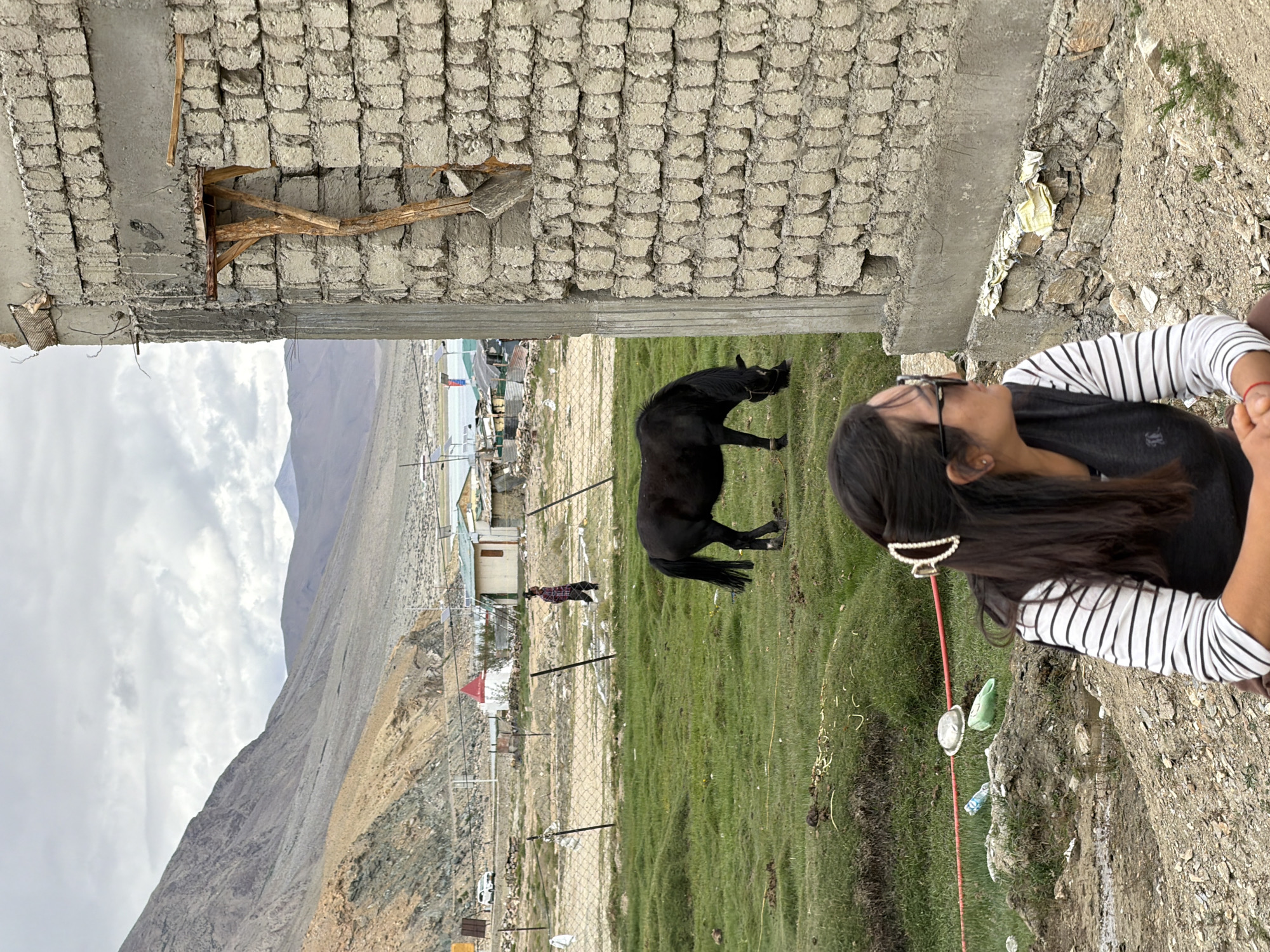 The image shows a young woman in the foreground, wearing a black vest over a striped long-sleeved shirt and dark sunglasses. She is looking to her right. Behind her, a black horse grazes in a grassy field. In the mid-ground, a person in a checkered shirt walks near a fence. A white car is parked in the distance, along with several buildings, some with red roofs. The background features a vast, mountainous landscape under a cloudy sky. A partially constructed brick building with exposed wooden supports is prominent in the right foreground. The scene appears to be in the high-altitude village of Karzok, India, located on the shores of Tso Moriri lake. The environment is a mix of natural landscapes and human settlement, with evidence of construction and scattered debris in the foreground.