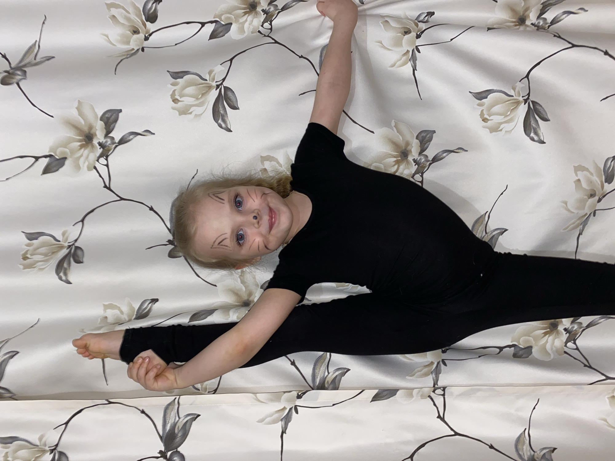 A young girl in Votkinsk, Russia, wearing a black leotard and leggings, lies in a split on a white floral-patterned fabric. She has fair skin, blonde hair, and blue eyes. There are drawn-on cat whiskers and a nose on her face. Her left arm is extended upwards, while her right hand is grasping her left foot.
