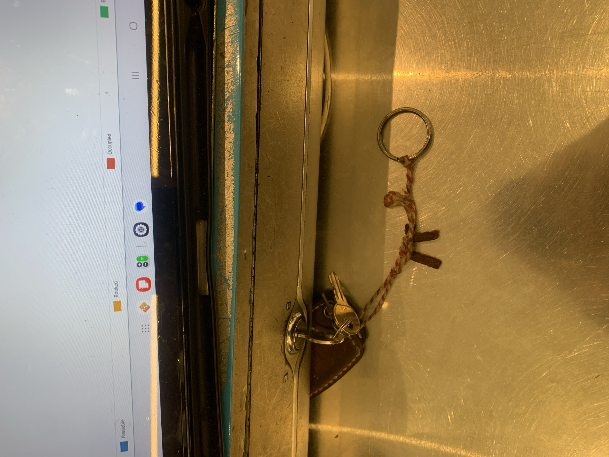 A photograph depicts a metal cabinet or container, possibly a safe or storage unit, with a lock and keys present. The keys are attached to a keyring and a small leather fob. Adjacent to the cabinet is the edge of a screen, possibly a tablet, displaying icons indicating availability and occupancy. The surface below the cabinet appears to be a brushed metal surface. There is no information in the image to suggest this is in North Kuta, Indonesia.
