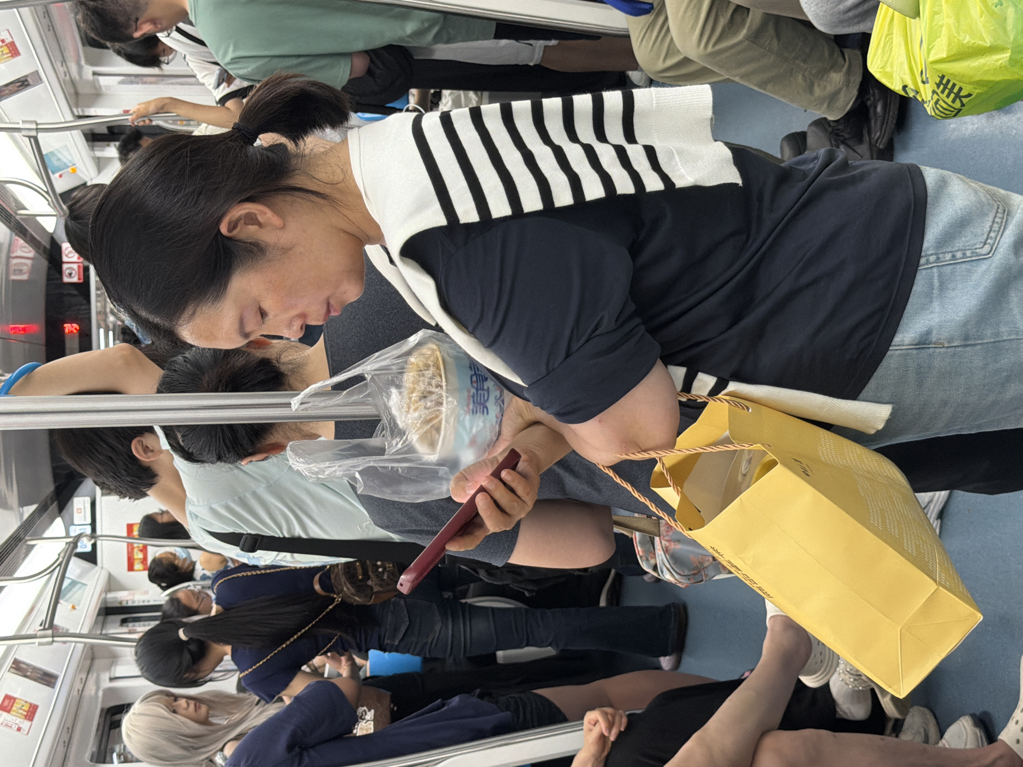 The interior of a crowded subway car in the city of Unknown, Unknown, is depicted under bright, artificial lighting. White walls, a white ceiling with silver overhead handrails, and a blue floor characterize the environment.
In the foreground, a woman with dark hair pulled back into a low ponytail is focused on a red smartphone held horizontally in both hands. She wears a dark blue short-sleeved t-shirt, light blue denim jeans, and has a white and black horizontally striped sweater draped over her shoulders. Her left hand also holds a clear plastic bag containing a white bowl of dry, light-colored food, which bears the Chinese characters "美美" (Mei Mei). A yellow paper shopping bag with braided rope handles hangs from her left wrist.
To the woman's immediate left, another woman with long, pale blonde hair stands facing away from the viewer, appearing to engage with a third woman with long dark hair tied in a high ponytail. The blonde-haired woman wears a dark blue top. The dark-haired woman wears a dark blue top and has a brown patterned backpack. Further into the background on the left, other indistinct figures are visible.
To the right of the foreground woman, several other passengers are present. One man in a light blue t-shirt has a black strap across his shoulder, and another man in a green t-shirt is partially visible further right. Many standing passengers are holding onto the overhead handrails. The floor in the foreground shows various footwear, including white sneakers and dark shoes. A partially visible bright yellow plastic bag with Chinese characters is on the floor to the far right.
