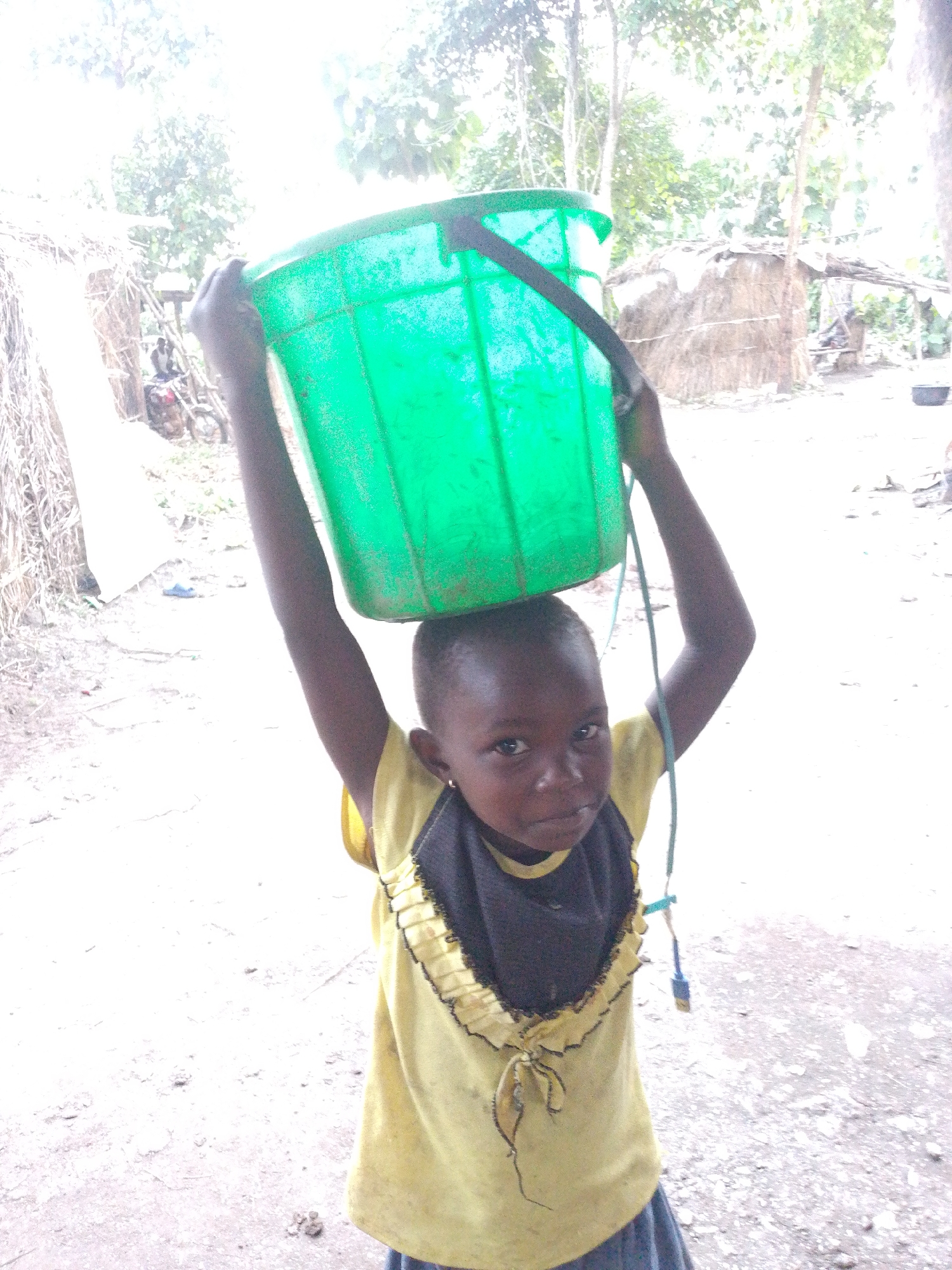 A young child, likely a girl given her attire, is carrying a large, bright green bucket of water balanced on her head. She is looking directly at the camera with a serious expression. The child is wearing a yellow t-shirt with a black ruffled neckline and a blue skirt. Her right arm is raised, supporting the bucket. A thin rope or strap hangs from the bucket, attached to a blue object.

The setting appears to be a rural or semi-rural area in Otulu, Nigeria. In the background, there are simple structures with thatched roofs, and dried vegetation. The ground is uneven and appears to be dirt or gravel. Trees and foliage are visible in the distance. The lighting suggests it is daytime, possibly overcast or with a strong backlight creating some lens flare. A person is visible in the background on a motorcycle. The overall scene depicts a child performing a common chore in the depicted location.