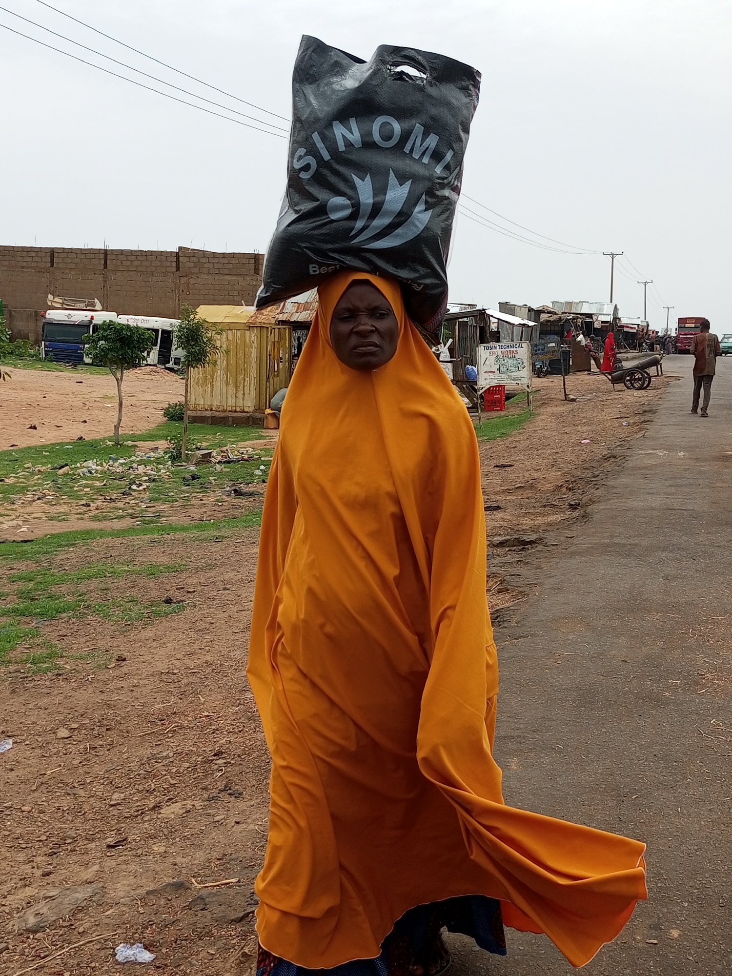 A woman, with dark skin and wearing a bright orange hijab and abaya, balances a large black plastic bag on her head. The bag has "SINOMIA" printed on it in white lettering, with a logo featuring curved lines. Her facial expression is serious, with her mouth slightly open and her eyes looking forward. She is walking on an asphalt road, with a dirt and grass embankment to her left. The embankment is littered with debris. In the background, there are several buildings with corrugated metal roofs, including what appears to be market stalls. A few people are visible in the distance, including a man walking away from the camera. There are also vehicles, including a truck, a bus, and a cart with wheels, parked or moving in the background. The sky is overcast. The scene appears to be in an urban or semi-urban setting, likely in the city of Gombe, Nigeria, given the context provided.