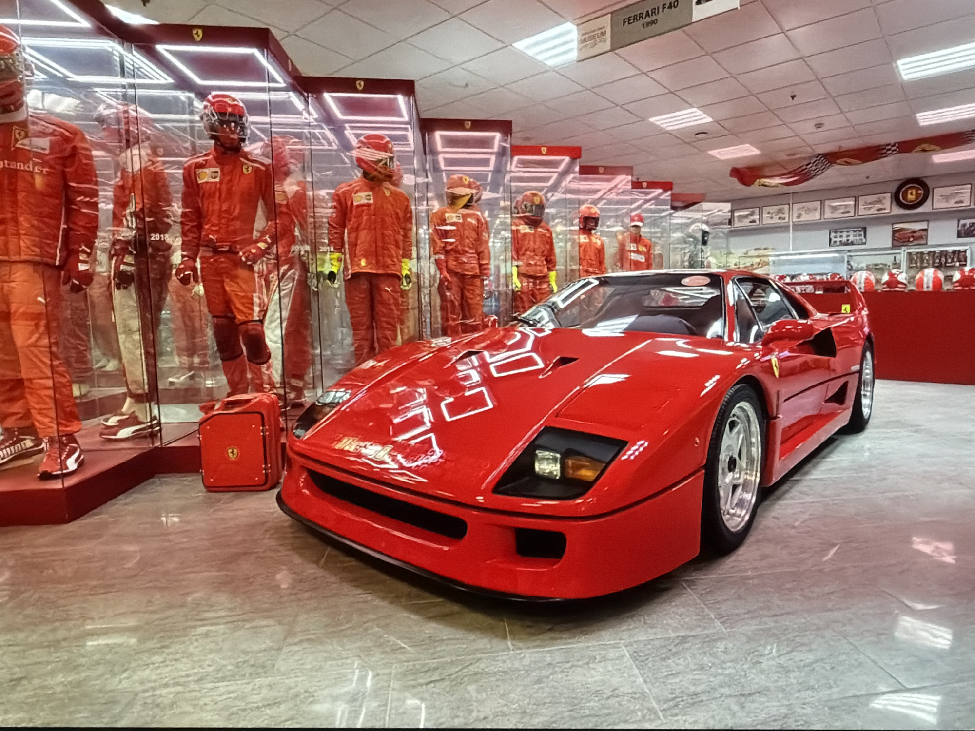 The image shows a red Ferrari F40 parked in a museum exhibit. Behind the car are several mannequins dressed in red racing suits and helmets, displayed within glass cases with illuminated red frames. Next to the first mannequin is a red gas can with a yellow Ferrari logo. In the background, there are framed pictures on the wall and a banner with a checkered pattern and the Ferrari logo. A sign above the exhibit reads "FERRARI F40 1990". The location is Unknown, Unknown.