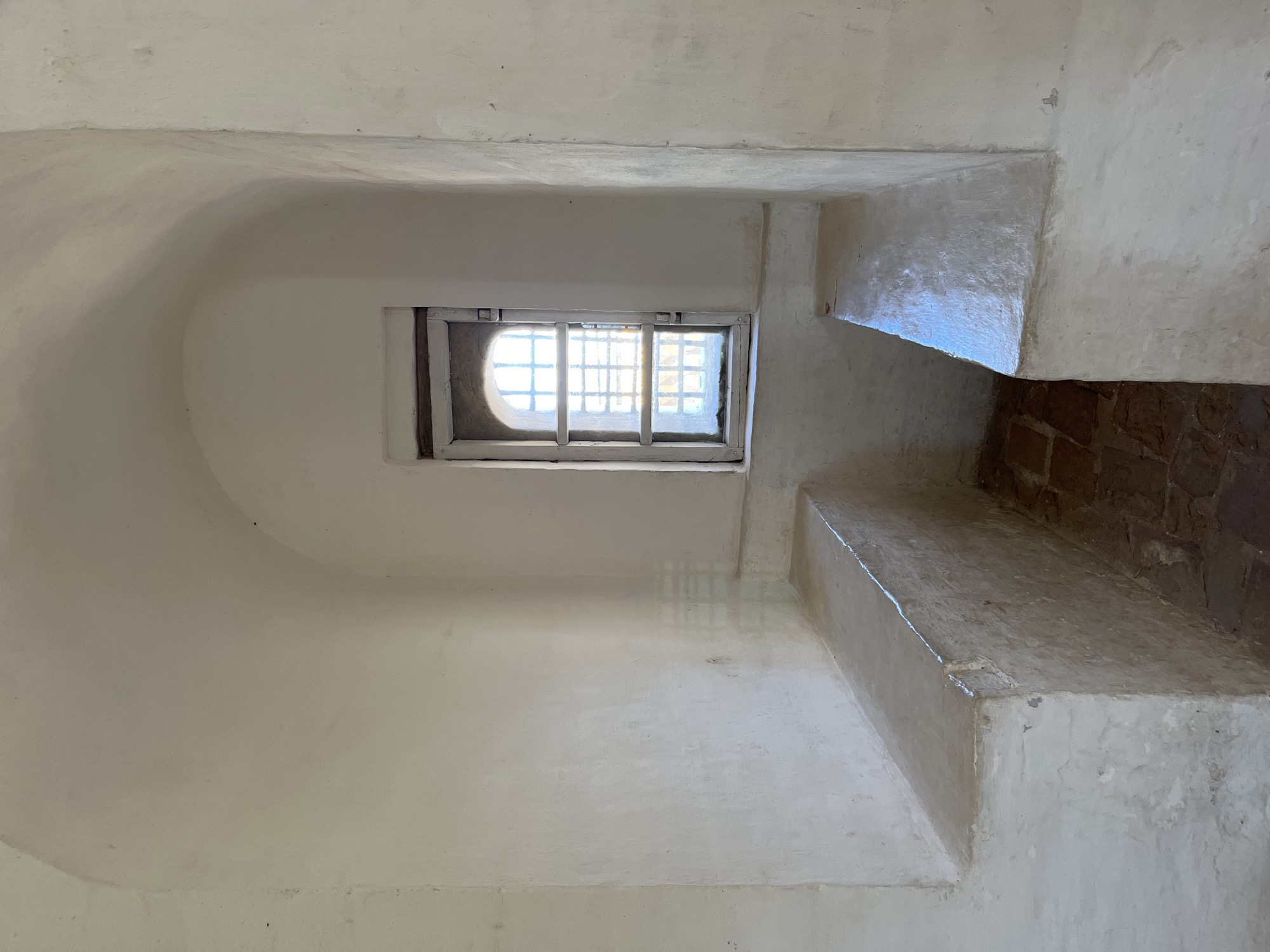 This is a photograph of the interior of a small, stark room with white, textured walls and a rounded ceiling. In the center of the back wall is a small, arched window with a wooden frame and metal bars. Sunlight streams through the window, casting striped shadows on the wall. On either side of the room, built into the walls, are two long, bench-like structures made of white plaster or concrete. The floor is covered with reddish-brown brick pavers. The scene appears to be inside a historical building, possibly a former prison cell or a similar austere space, located in Kladovitsy, Russia. There are no people or animals visible in the image.