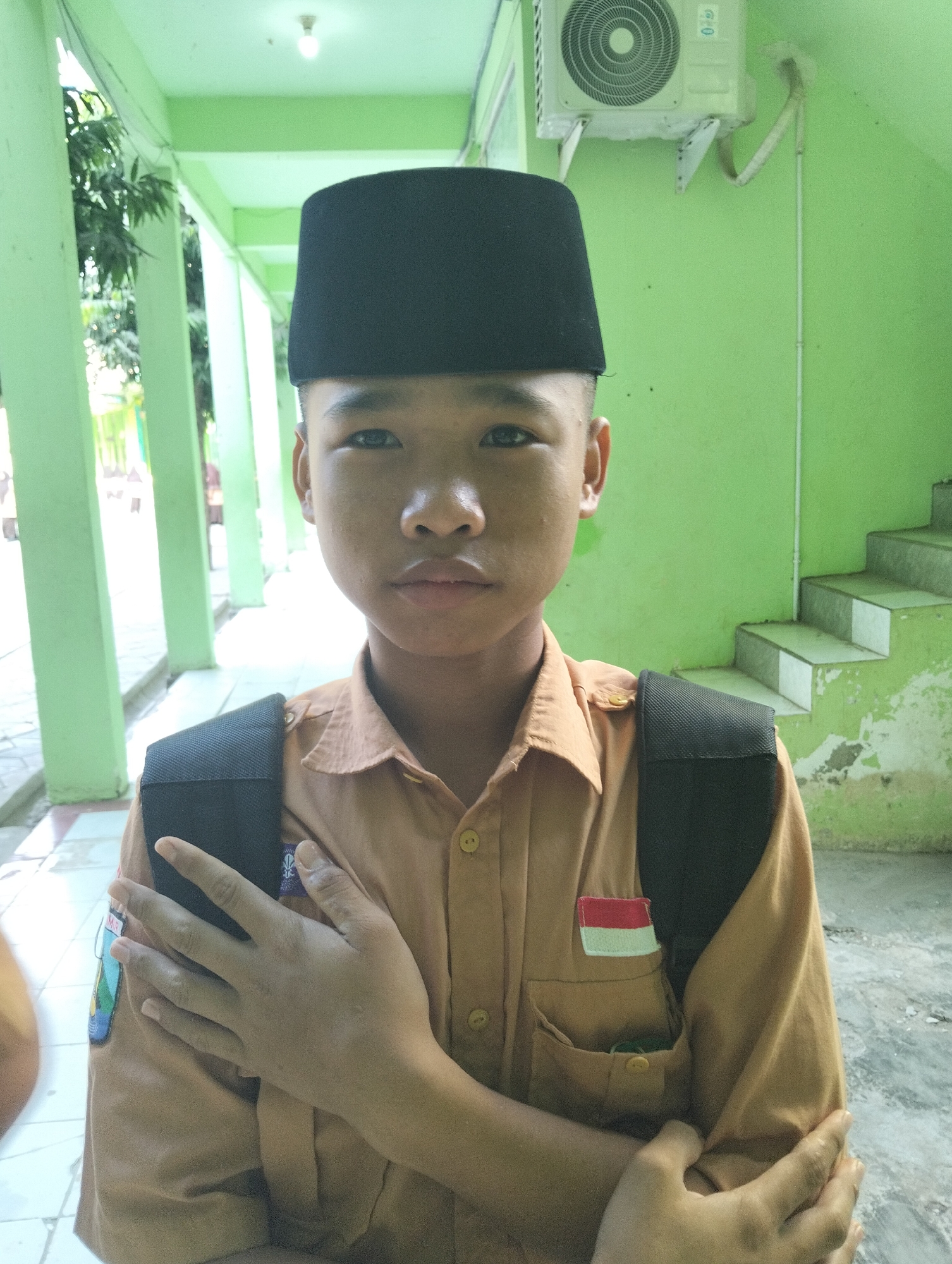 A young Indonesian boy is positioned centrally in the frame, facing forward. He wears a dark peci hat and a light brown uniform shirt with a backpack strap visible over his shoulders. A small Indonesian flag patch is affixed to his shirt pocket. His hands are crossed in front of his chest. The background appears to be an outdoor hallway or covered walkway with light green painted walls and pillars. In the upper right corner, an air conditioning unit is mounted on the wall. To the right, a set of concrete stairs ascends against the green wall. The ground is paved with a light-colored material. The lighting suggests it is daytime, with ambient light illuminating the scene. The overall environment seems to be an educational institution, possibly a school in Paciran, Indonesia, given the context of the uniform and the architectural style.