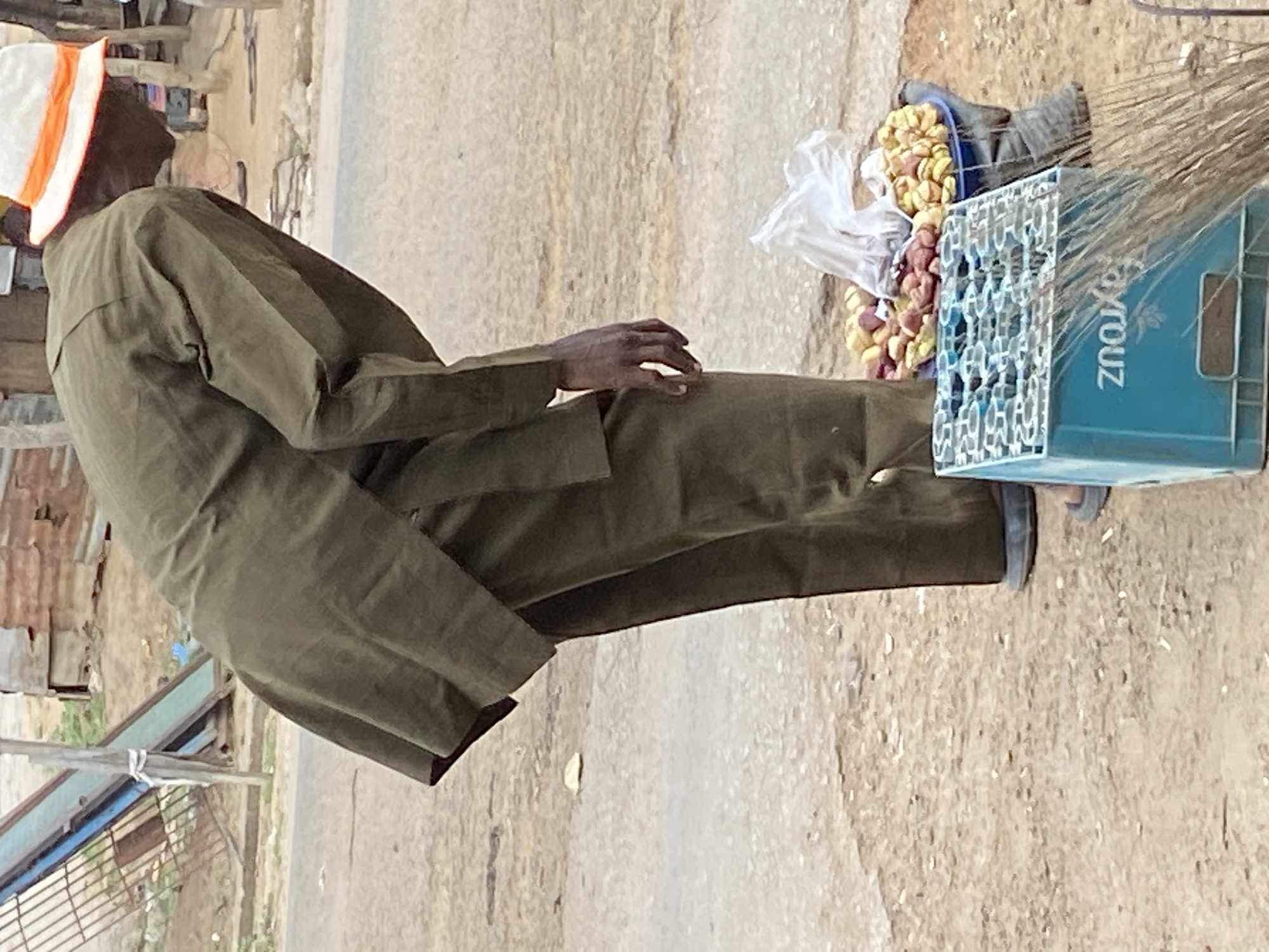 The image shows a man in Mararaba, Nigeria, sitting on the side of a road. He is wearing a long-sleeved green outfit and a white hat with orange stripes. Next to him are fruits, possibly groundnuts, packed in a bag and a bucket. A blue plastic crate is situated beside the fruits. A broom is leaning against the crate. The background consists of a sandy and rocky roadside. A partially visible fence and buildings are in the distance.