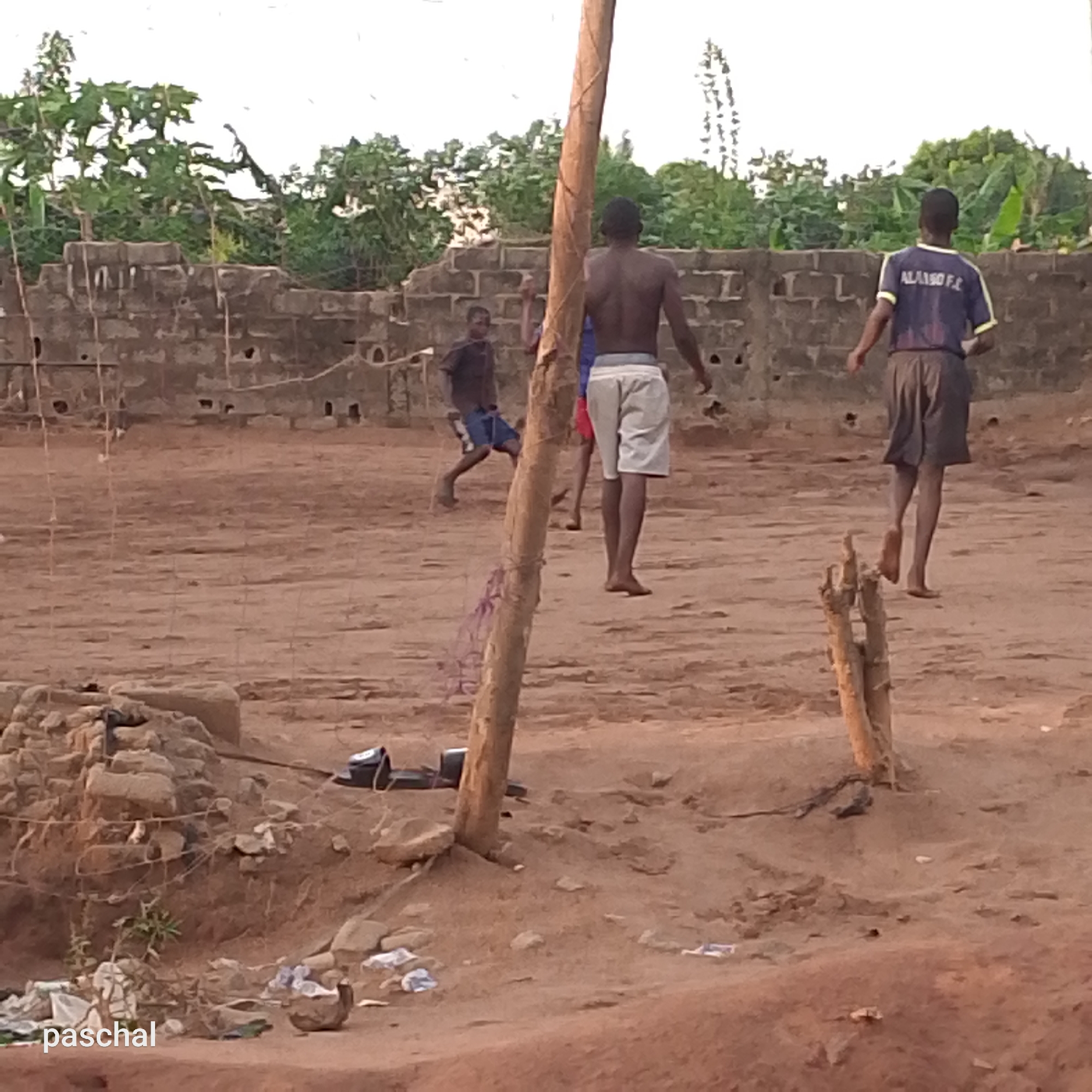 The image depicts an outdoor scene in Ukwu-Abwa, Nigeria, featuring a group of young men playing a game on a dirt field. A dilapidated brick wall borders the field, with overgrown vegetation visible behind it. Several wooden posts are positioned in the foreground, possibly delineating a boundary or goal. The ground is bare dirt, littered with small objects and debris. Three of the players are more clearly visible: one is shirtless and wears light-colored shorts, another wears a dark blue jersey with "ALABO F.C." printed on the back, and the third wears a dark shirt with blue shorts. They appear to be playing barefoot or in minimal footwear. A pair of dark sandals lies on the ground near one of the wooden posts. The lighting suggests it might be late afternoon or early morning, with a slight reddish hue to the scene.