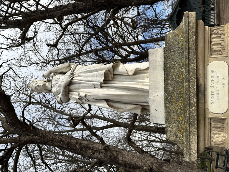 The image displays a weathered white marble statue of Sainte Bathilde, Queen of France, situated outdoors in a public park, specifically the Jardin du Luxembourg in Paris, France. The statue, depicting a regal female figure, stands prominently on a multi-tiered stone pedestal.
The figure wears a crown and long, flowing robes with intricate draping. Her left arm is bent, holding a vertical, staff-like object, while her right hand rests over her left forearm. Her gaze is directed slightly upwards and to the viewer's left. The statue exhibits subtle discoloration and weathering consistent with prolonged outdoor exposure.
The stone pedestal beneath the statue is rectangular and shows patches of greenish-yellow lichen and moss, particularly on its upper surface. A white plaque embedded in the front of the pedestal bears the inscription "SAINTE BATHILDE / REINE DE FRANCE / - 600" in black capital letters. Decorative relief carvings are visible on the lower section of the pedestal.
The background consists primarily of a dense network of bare tree branches against a light, overcast sky, indicating a winter or early spring season. The trees feature thick, dark trunks and gnarled branches. To the far right, beyond the trees, a dark green, hexagonal or octagonal gazebo-like structure with a pointed roof and white trim is partially visible. A dark green park bench is partially visible in the lower-left corner of the frame, near the base of a tree. No people are present in the scene.