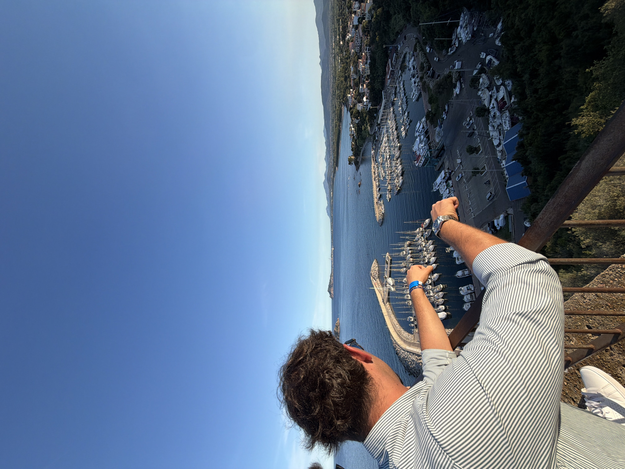 An elevated, over-the-shoulder perspective looks out from a balcony at a man facing away from the camera, his head and shoulders in the foreground. He wears a light-colored, horizontally striped shirt and white sneakers. His right arm is extended, resting on a rusty railing. The man is looking towards a large harbor filled with numerous sailboats and smaller vessels.
The harbor is situated in Santa Maria Navarrese, Italy, and is surrounded by a coastal town with buildings clustered along the shoreline and extending inland. A long, curved breakwater protects the harbor. Beyond the town and harbor, a verdant landscape with hills and mountains forms the backdrop under a clear, pale blue sky. The scene is bathed in the warm light of late afternoon or early evening.