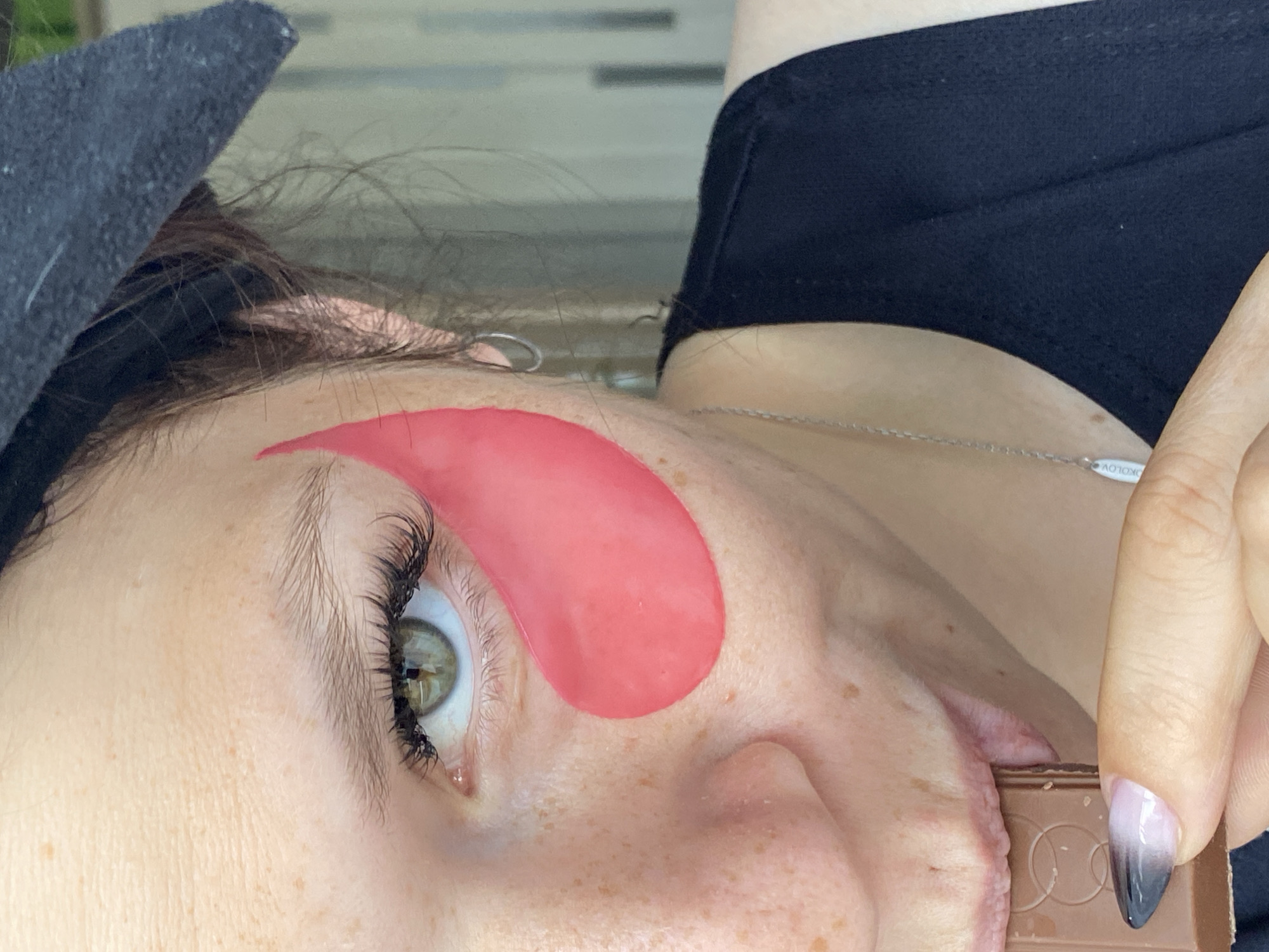 A close-up shot shows a woman with fair skin, freckles, and green eyes. She is wearing a black headband, a black top, and a silver necklace. Her left eye is adorned with a pink under-eye patch. Her nails are painted with a gradient of black and pink. She is holding a piece of chocolate and biting into it. The background is blurred and appears to be indoors, with vertical elements suggesting blinds or a window. There is no identifiable city or location information present in the image.