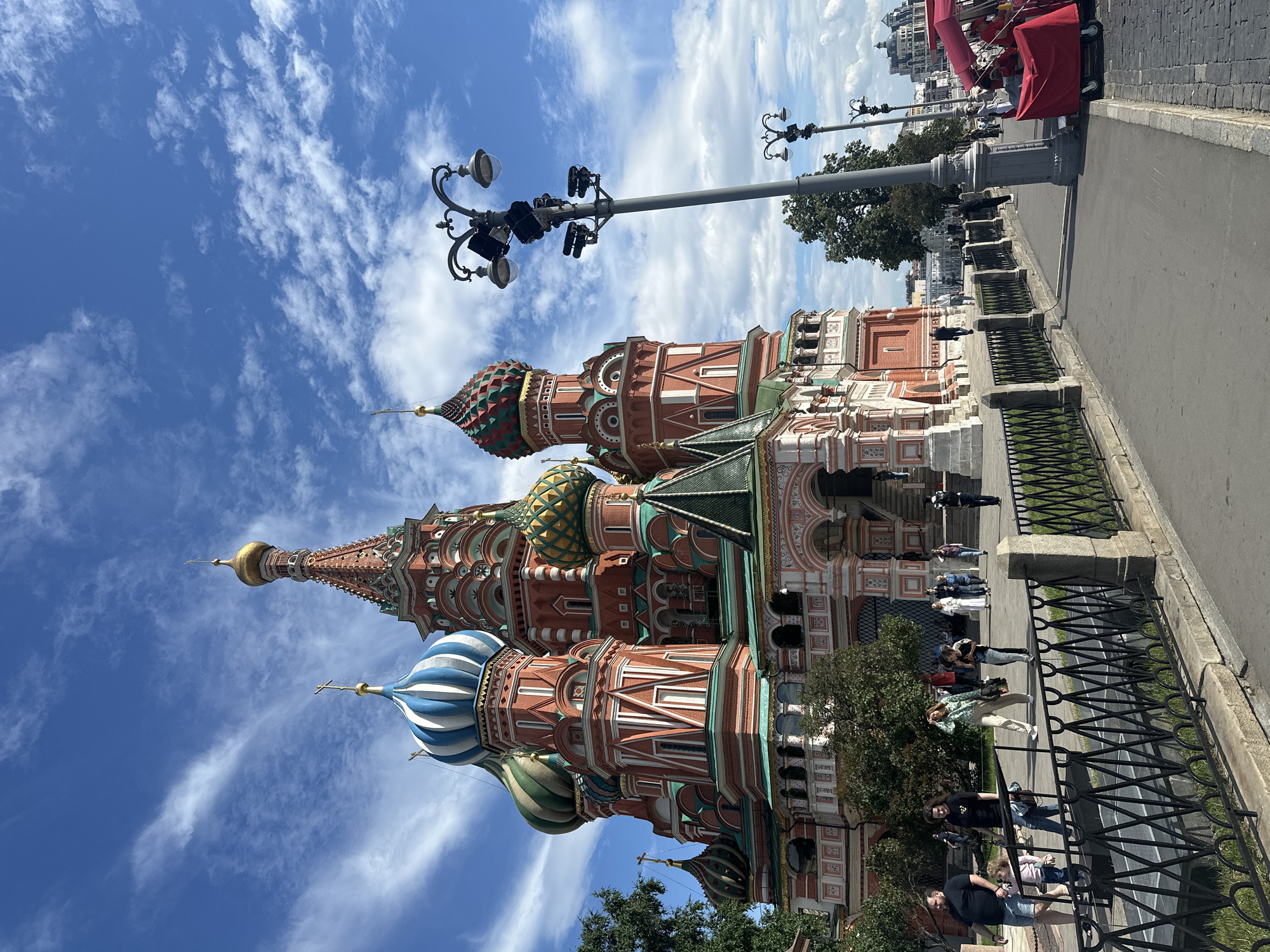 The image shows Saint Basil's Cathedral in Moscow, Russia, on a sunny day with scattered clouds. The iconic cathedral, with its vibrant red and white striped facade and colorful onion domes, dominates the center and left side of the frame. A black metal fence runs along the foreground, separating a paved walkway from a grassy area. People are walking on the walkway, some singly and others in small groups. To the right, a lamppost with multiple lights stands tall, and further down the walkway, a red cart with a canopy is visible. In the background, other buildings and trees can be seen.