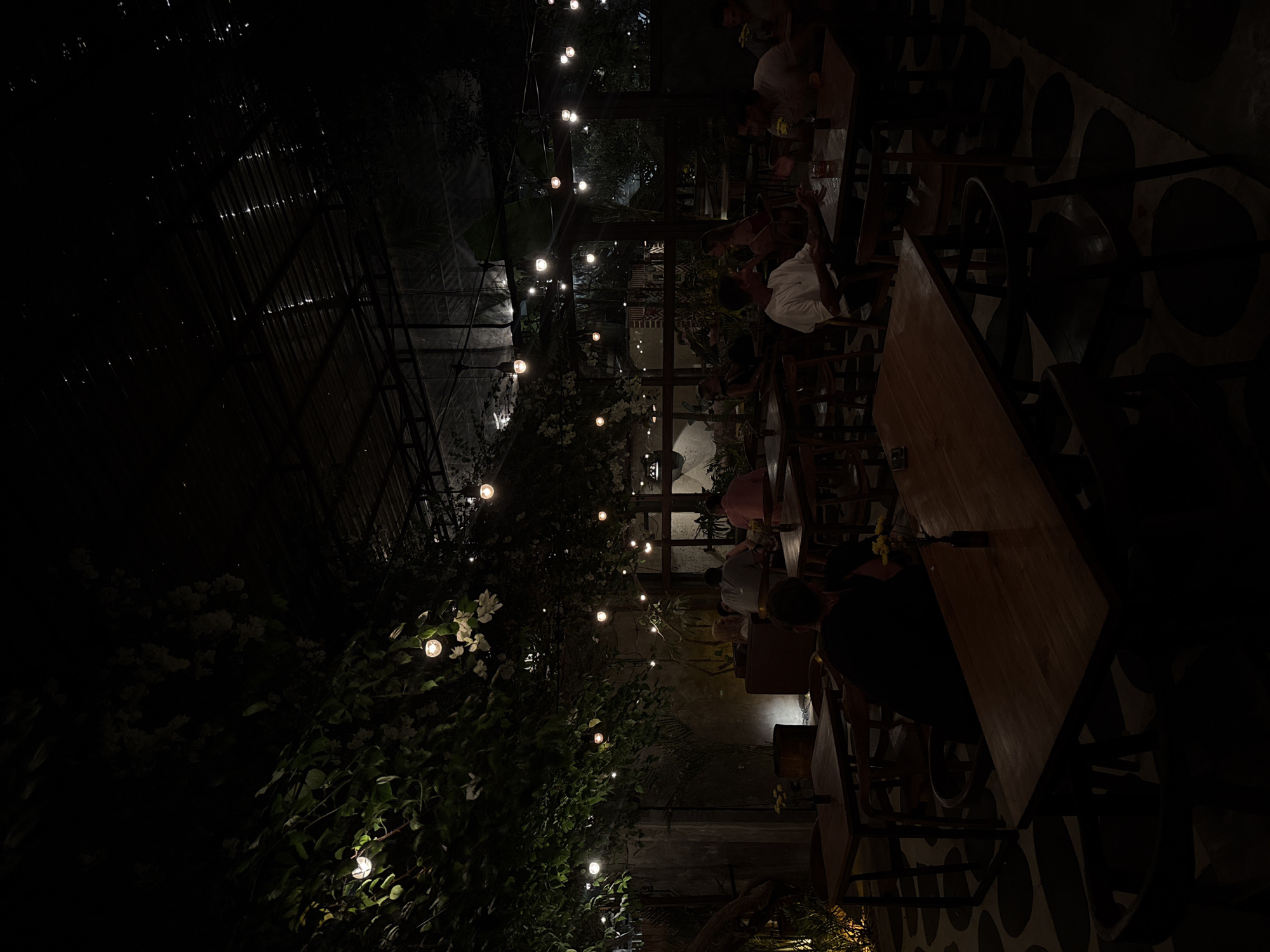 The image captures an outdoor dining area at night in Kuta Selatan, Indonesia. Several tables are occupied by people. String lights are strung above, illuminating the area, particularly through foliage. Some of the overhead structure appears to be a greenhouse. The floor has a patterned design. People are seated at the tables, seemingly enjoying a meal or drinks. The overall scene suggests a relaxed and ambient atmosphere.