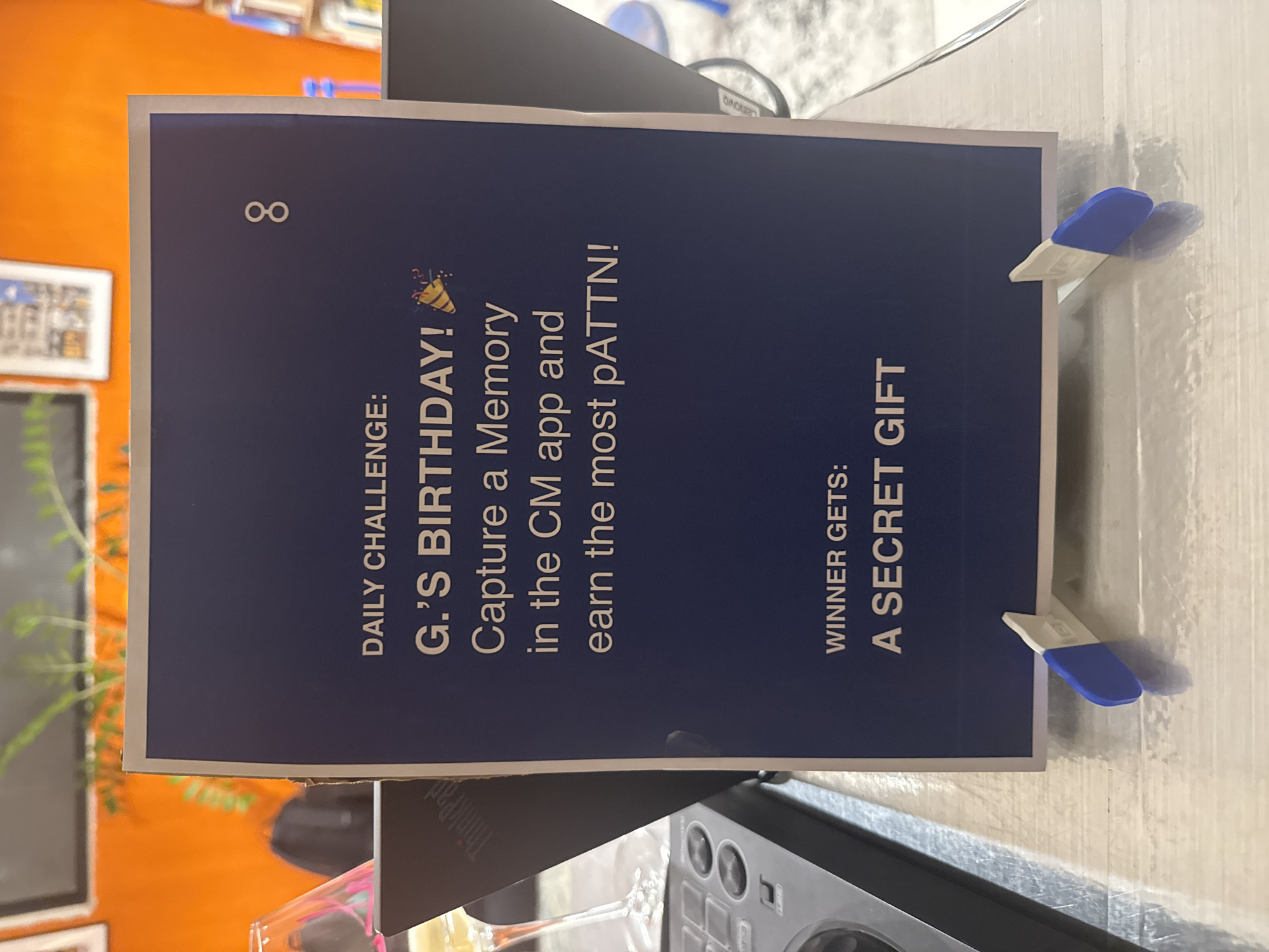 A navy blue sign with white text stands upright on a table, supported by two small blue and white stands. The sign announces a "DAILY CHALLENGE: G.'S BIRTHDAY! Capture a Memory in the CM app and earn the most pATTN!". Below this, it states "WINNER GETS: A SECRET GIFT". A single number "8" is printed in white in the upper right corner of the sign. In the background, a person's arm holding a champagne flute is visible, and behind them, a brightly lit orange wall is present. The scene appears to be indoors, possibly in a home or office environment, and there is no specific indication of a location within Tel-Aviv, Israel.