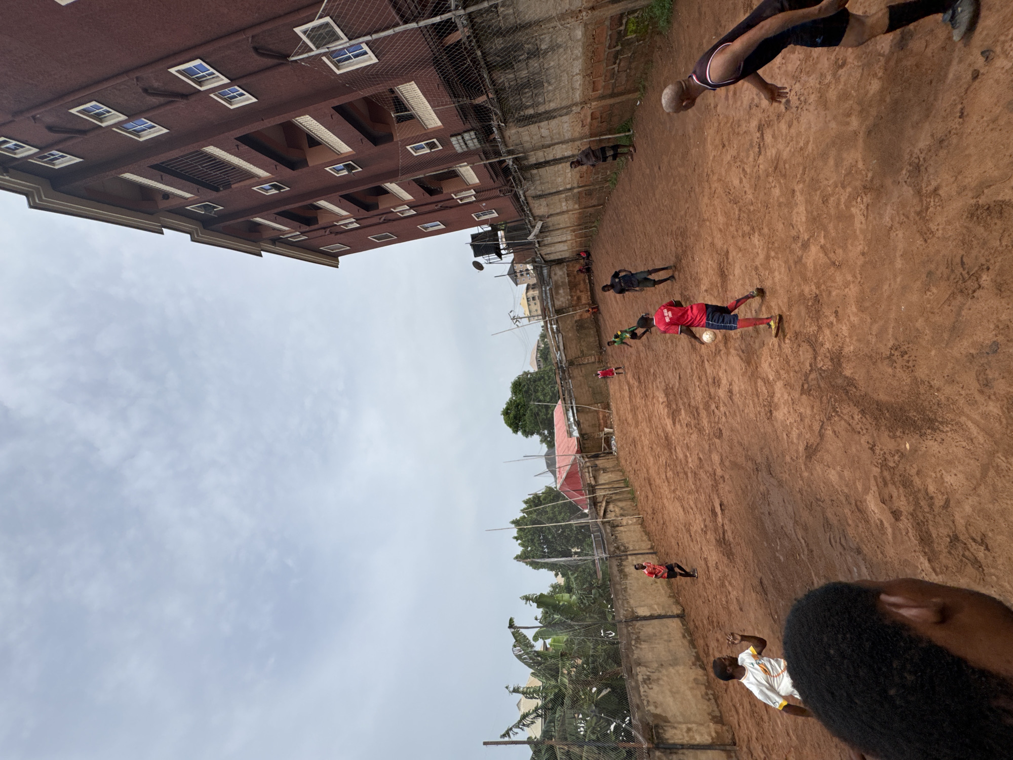 The image shows a soccer game in progress on a red dirt field in Nkpor, Nigeria. Several players in red and other colored shirts are actively involved in the game. Some people are standing or moving along the periphery of the field. The backdrop consists of a building and a wall with vegetation. The sky is overcast.