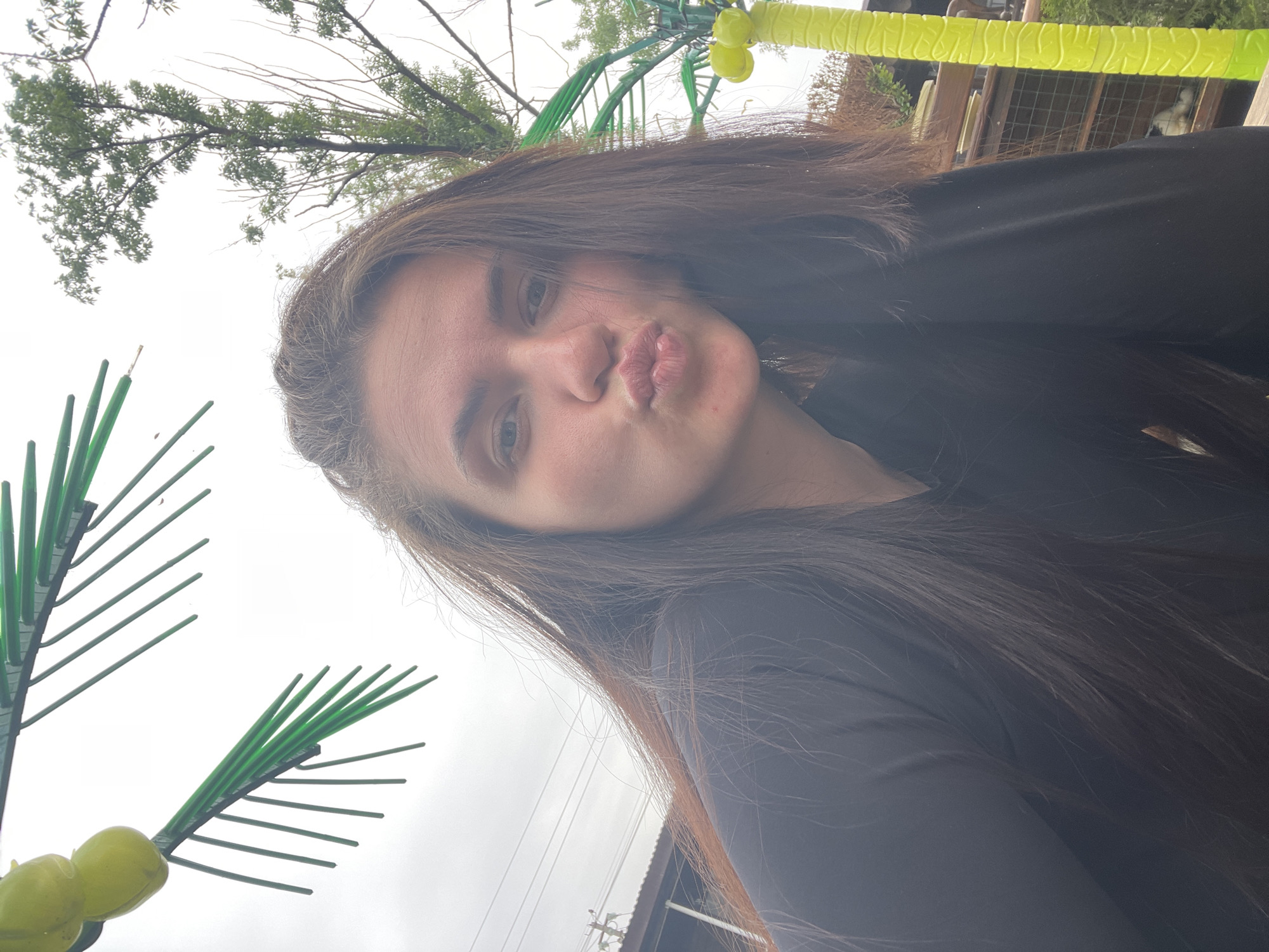 A young woman with long brown hair, wearing a black long-sleeved shirt, is making a kissing face toward the camera. She appears to be outside, with a cloudy sky visible in the background. To the left of the frame is a green plastic palm tree, and above her, partially obscured by her hair, is a real tree with foliage. A fence and a partial view of a building structure are also visible in the background. The location is Molokovo, Russia.