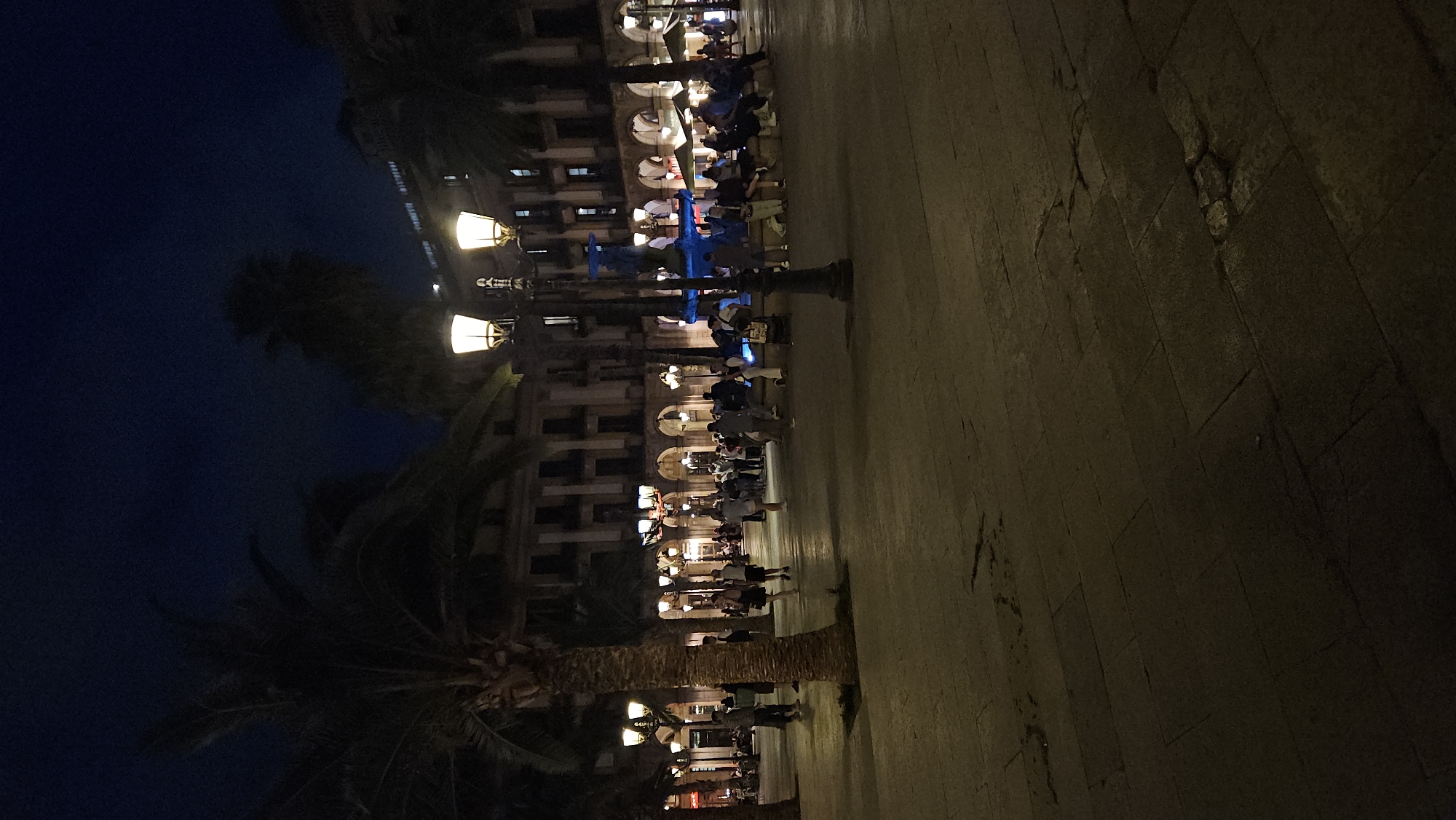 The image displays Plaça Reial in Barcelona, Spain, at night. The scene is illuminated by numerous artificial light sources, primarily warm-toned electric lampposts. The sky is a deep, dark blue, indicating late evening or nighttime. The ground consists of a textured pavement of large, rectangular stone tiles. Several tall palm trees are strategically placed throughout the square, their dark fronds silhouetted against the ambient light. Surrounding the plaza are multi-story buildings featuring arcaded ground floors with numerous arches, some housing outdoor cafe or restaurant areas. A distinctive, multi-tiered fountain, painted blue, is centrally located in the midground, with people seated around its base. A prominent, ornate black lamppost with three glowing lamps stands in the foreground, consistent with Antoni Gaudí's designs for Plaça Reial. Numerous individuals are present in the square; some are walking across the paved area, while others are standing, conversing in groups, or seated on benches around the fountain and at tables under the building arcades. The overall environment suggests a lively public space.