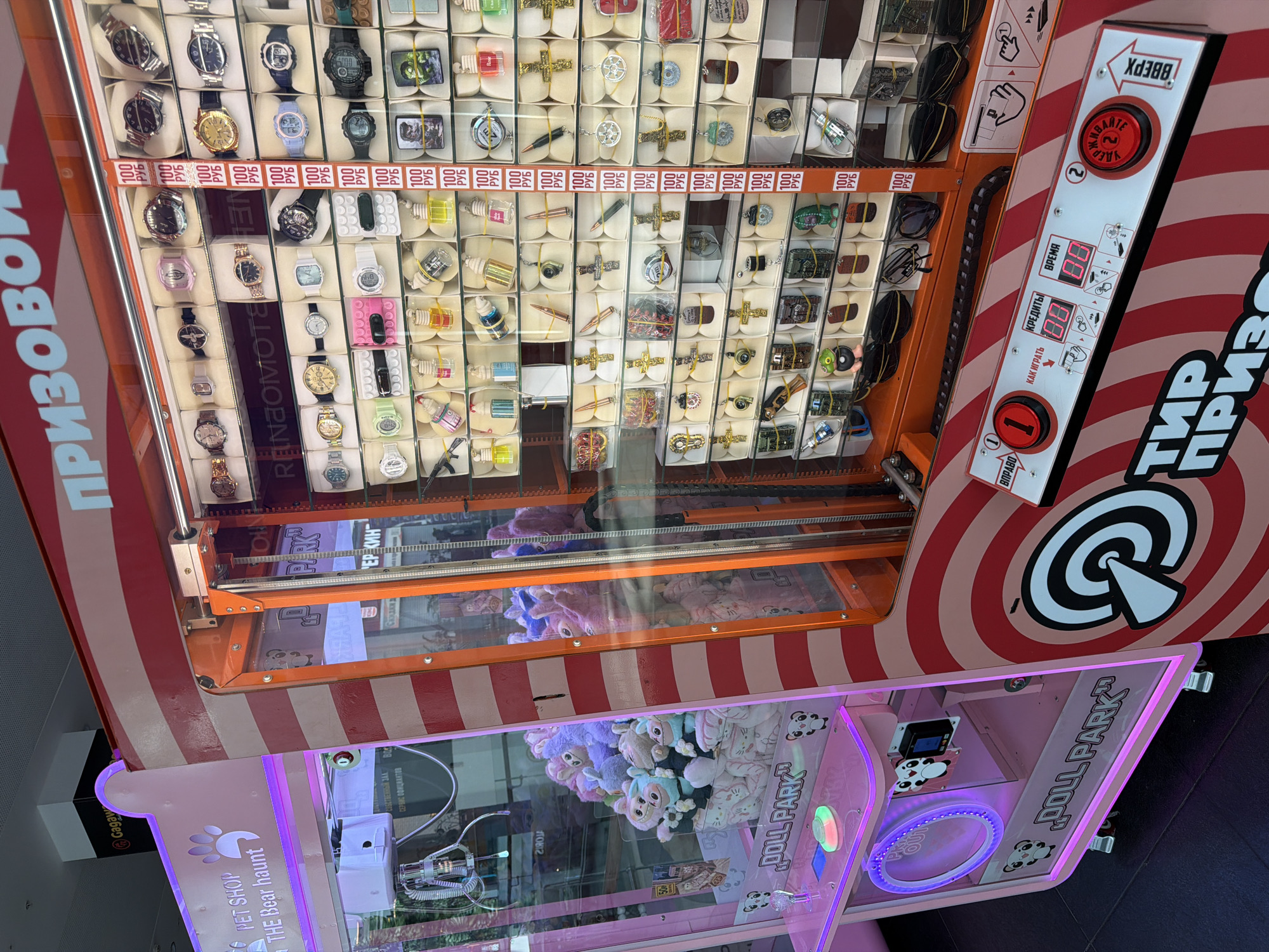 This image displays two arcade game machines located in an indoor entertainment venue, likely in Moscow, Russia. No people are visible in the frame, but the machines are brightly lit and appear ready for interaction.
The upper machine, with an orange and red striped design, is a prize game identified by the Cyrillic text "ТИР ПРИЗОВ" (Prize Shooting Range/Gallery) and "ПРИЗОВОЙ" (Prize). Its transparent front showcases numerous small prizes organized in individual compartments, including various styles of watches (both digital and analog), keychains, and small gadgets. Each compartment features a "100 РУБ" (100 Rubles) price tag. A white control panel is equipped with red buttons labeled "ВПРАВО" (Right), "ВВЕРХ" (Up), and a prominent "ЖМИТЕ УДЕРЖ" (Press Hold) button. Digital displays show "КРЕДИТЫ" (Credits) and "ВРЕМЯ" (Time) both set to "00," along with instructions on "КАК ИГРАТЬ" (How to Play).
Below it, a pink claw machine is adorned with purple LED lighting and branded with "DOLL PARK" and "PET SHOP THE Bear haunt." Inside, a large collection of soft plush toys, including pink, purple, and blue stuffed animals, fills the playing area. A metallic claw mechanism is visible, positioned above the prizes.
Reflections of the surrounding environment, such as other commercial signage and elements like "Gagaw" and "СЕРВИС ОФИЦИАНТОВ" (Waiters service), are faintly visible on the glass panels of both machines, suggesting a larger, bustling indoor setting. The scene conveys an atmosphere of recreational gaming and prize-winning.