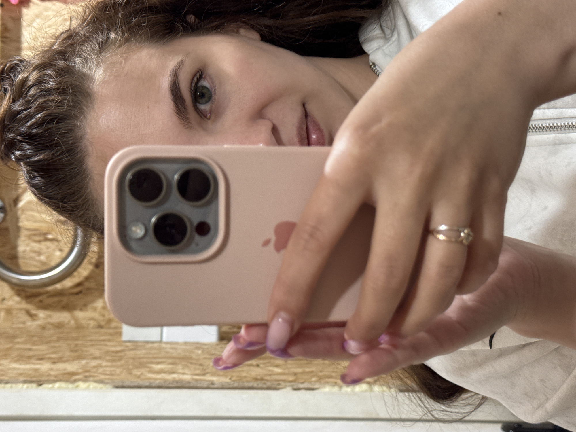 A woman is taking a selfie with a pink iPhone. She is wearing a white shirt. She has a ring on her finger. The location appears to be a bathroom, with a wooden wall and white trim. The location of Nizhnyaya Gar', Russia cannot be verified from the image.