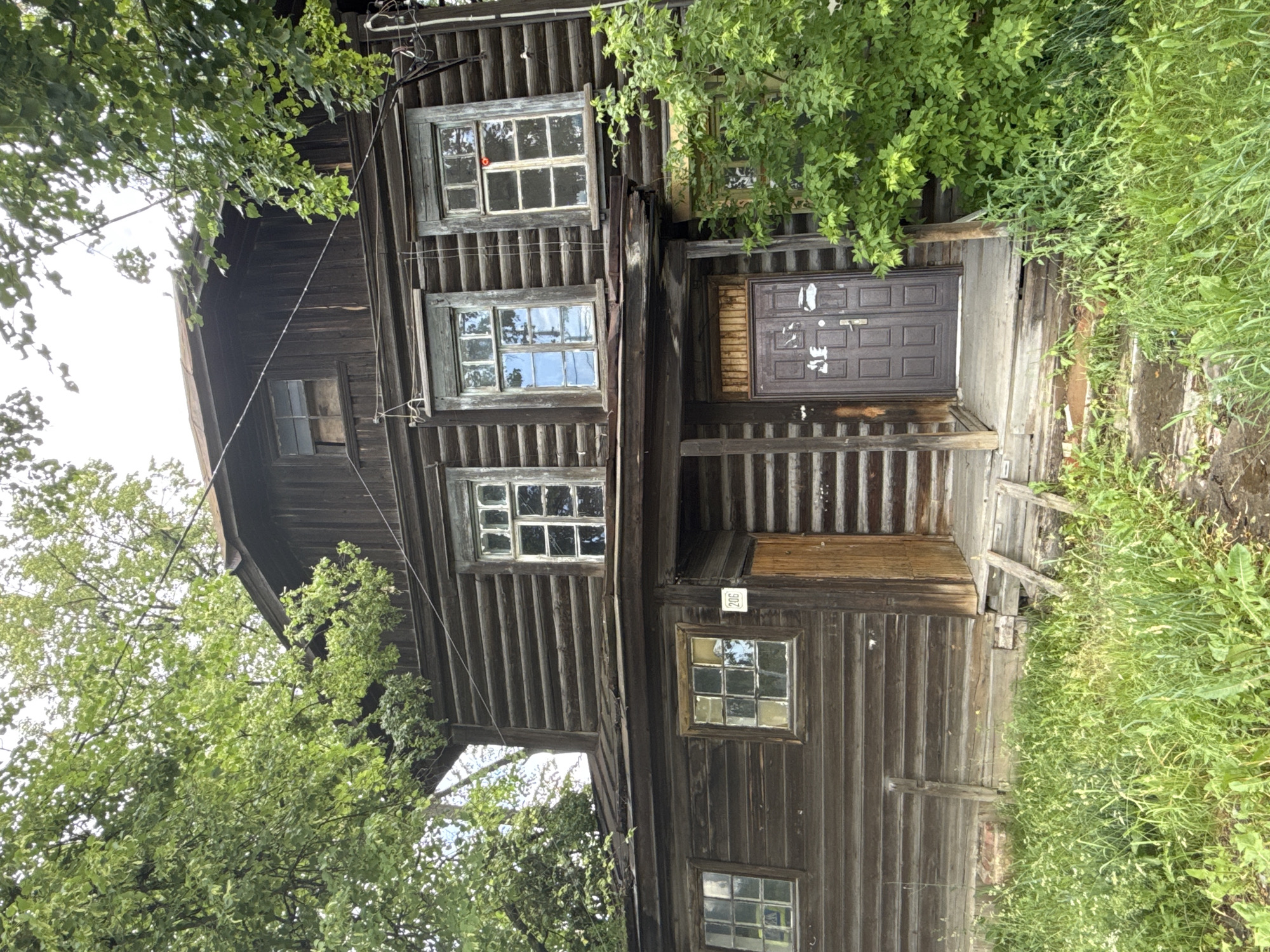 The image depicts the exterior of a weathered, two-story wooden log house located in the city of Izhevsk, Russia. The house is surrounded by lush green foliage and tall trees, with overgrown grass and weeds in the foreground. The wooden walls of the house show signs of age and wear. There are several windows on both floors, some with glass panes that appear dusty or partially obscured. A porch with a wooden door is visible at the front of the house, with a small number plate reading "206" above a wooden door to the left. The overall impression is one of an old, possibly abandoned or neglected, rural dwelling within an urban setting.