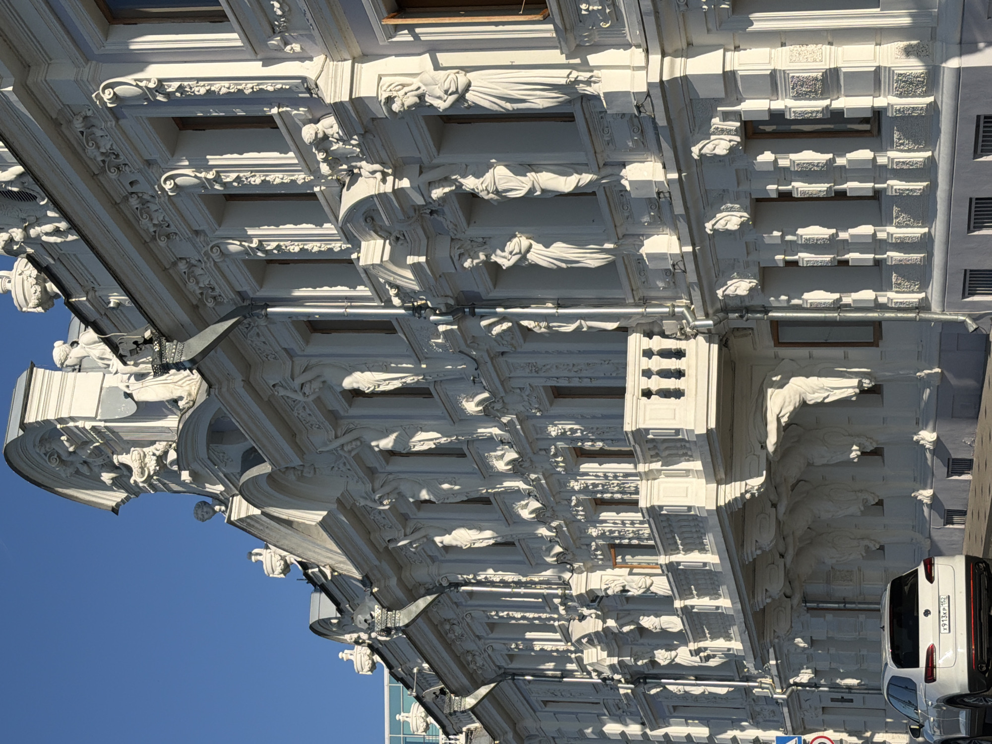 This image shows a close-up, exterior, street-level view of a white, ornate, multi-story building in Nizhny Novgorod, Russia, bathed in sunlight. The architecture is highly decorative, featuring numerous sculptures of female figures, ornate carvings, and elaborate cornices and window frames. The building appears to be historical, with a classical style. The sky is a clear, bright blue.
In the foreground, a white SUV is parked on the paved street in front of the building. A blue and white street sign is visible on the left side of the frame, indicating a traffic regulation. The building's facade is rich with details, including balconies supported by atlantes, statues in niches between windows, and detailed friezes. The windows are tall and narrow, with dark frames. The lower portion of the building has a plainer facade with barred windows. The sunlight casts strong shadows, highlighting the three-dimensional nature of the architectural embellishments. The overall impression is one of grandeur and historical significance.
