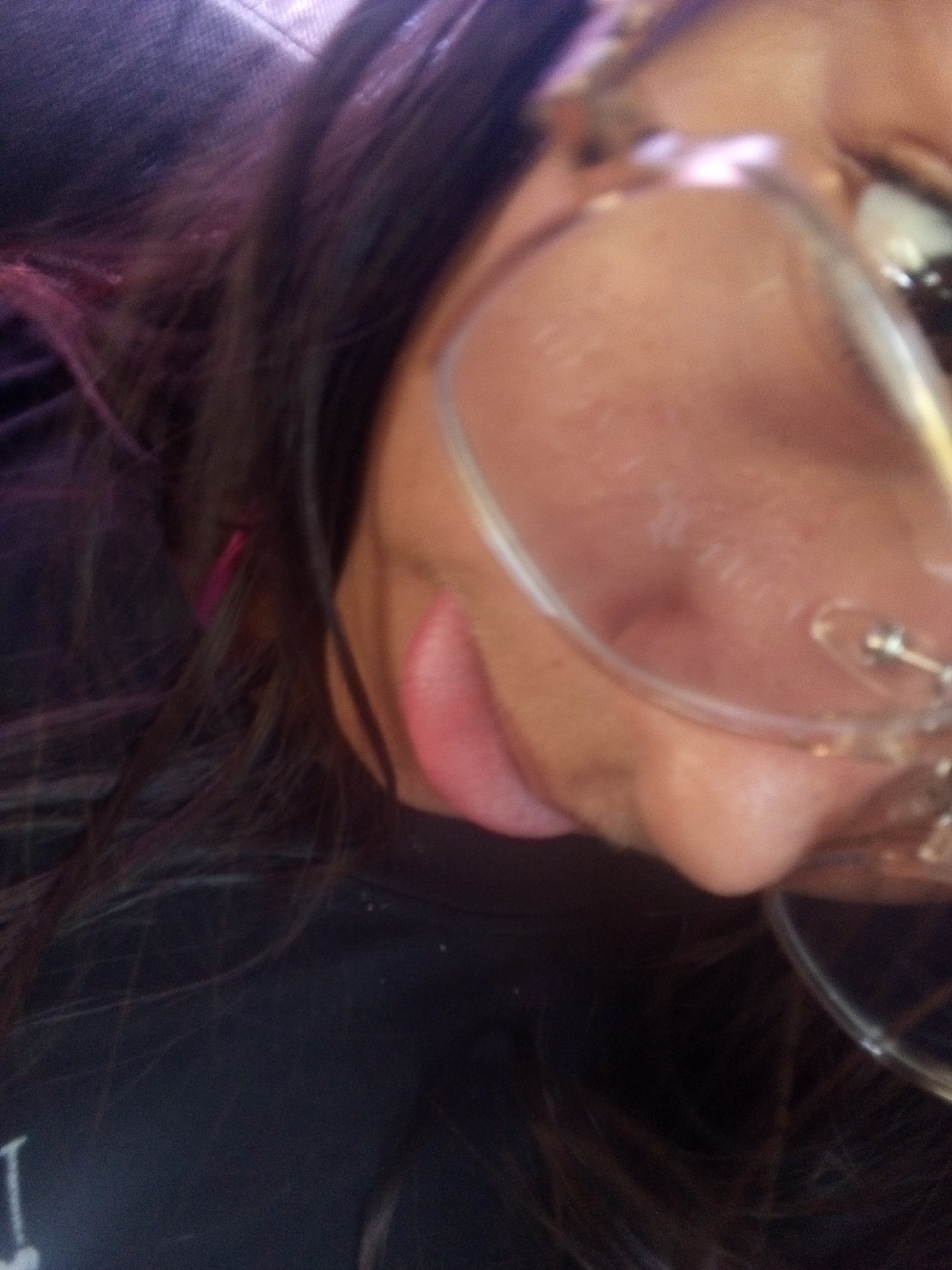 A close-up, angled photograph shows a person's face, partially obscured by eyeglasses. The individual has dark hair, with some strands showing a pinkish tint, and their skin appears to be fair. The person's lips are visible, and part of their neck is also in view. They are wearing a dark-colored shirt. The background is blurry and indistinct. There is no information in the image to determine the location of the image in Juan Griego, Venezuela.