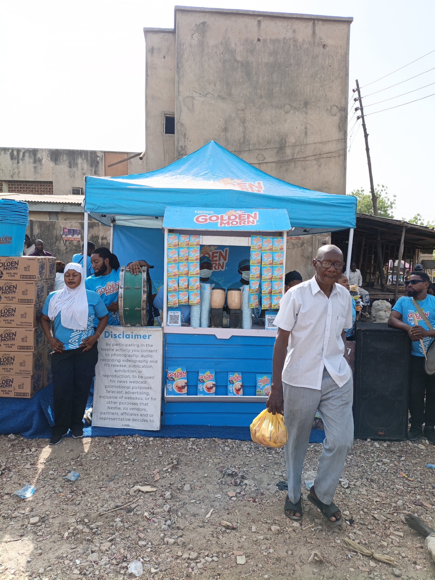 Here is a description of the image:
The image shows a "Golden Morn" promotional stand in what appears to be a street scene, likely in the city of Kawaji, Nigeria. The stand is predominantly blue and features the "Golden Morn" brand logo and cereal packaging.
Several people are visible. There is a group of people wearing matching "Golden Morn" t-shirts staffing the stand. A woman in a white headscarf is near the stand. An older man is walking in front of the stand, carrying a yellow bag.
The environment is somewhat cluttered. Some boxes of "Golden Morn" cereal are stacked next to the booth. The ground appears to be a dirt road. There are some simple buildings and an old power pole in the background. A large black speaker is near the booth, and a man in sunglasses is standing nearby with a drum.