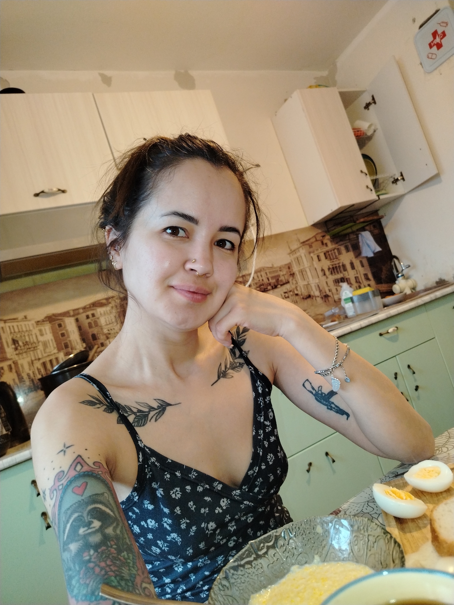 A woman with tattoos is sitting at a table in a kitchen. She has her hair pulled back and is wearing a black and white floral print dress. She is resting her chin on her hand. On the table in front of her are a bowl of food, sliced hard-boiled eggs, and bread. Behind her are white cabinets and a backsplash with a cityscape print. A first aid kit is visible on the wall. The kitchen appears to be in a home. There is no verifiable information regarding the city of Noviy, Russia.