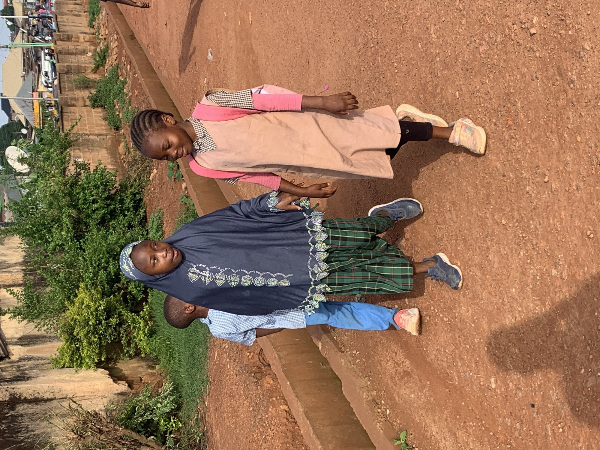This media file depicts a group of three young individuals outdoors. The scene appears to be a street or pathway with a reddish-brown, dirt ground and some vegetation along the side. In the background, there are buildings and what looks like a satellite dish.
The main subjects are three children. The eldest, a girl wearing a dark blue hijab and a patterned dress, stands in the center. To her left, a younger child in a light pink dress with a checkered undershirt is visible from the waist down, with their legs and feet showing. To the right of the eldest child, another young child wearing a blue checkered shirt and blue pants is also visible from the waist down. The children are standing, and their posture suggests they might be posing for a photograph or interacting with each other.
The sunlight casts shadows on the ground, indicating it is likely daytime. The weather appears to be clear. The environment suggests a warm climate, possibly a rural or semi-urban area. The children's faces are not clearly visible, so it's difficult to ascertain their emotions.
No visible text is present in the image.
