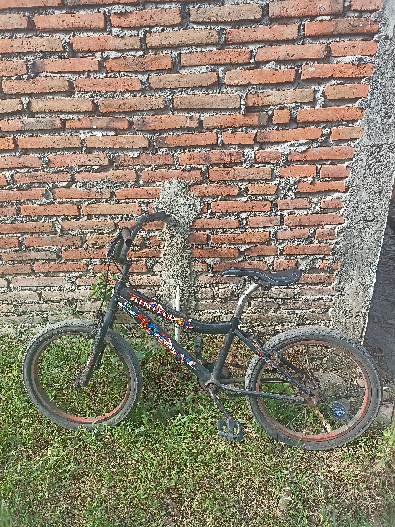 A black bicycle is leaning against a brick wall in a grassy area. The bicycle has red rims on its wheels and appears to be a BMX bike, with "UNITED" printed on the frame along with various superhero decals. The brick wall is weathered and shows signs of age. The ground is covered in green grass and some dry vegetation. A concrete pillar or structure is visible on the right side of the wall. The scene appears to be outdoors, likely in a residential area, possibly in Mojopanggung, Indonesia, given the context. There are no people or animals visible in the image.