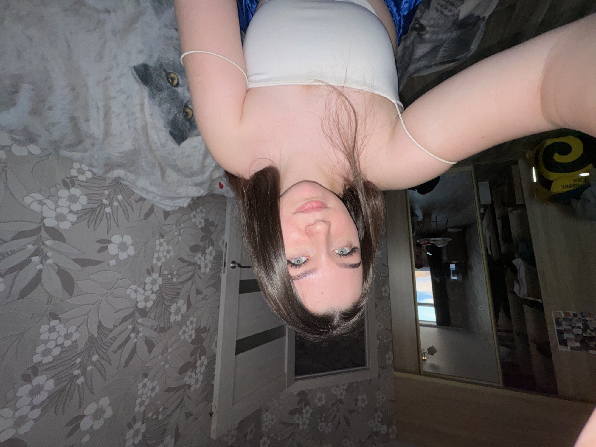 A young woman with long, dark brown hair and light green eyes is taking a selfie in what appears to be a bedroom. She is wearing a white strapless top and blue bottoms. The room has floral wallpaper and a large mirror reflecting another room with a window. There is a yellow bag with a black spiral design on the left side of the image, and a blanket with a cat pattern on the right. The scene appears to be indoors, possibly in an apartment in the city of Gamovo, Russia.